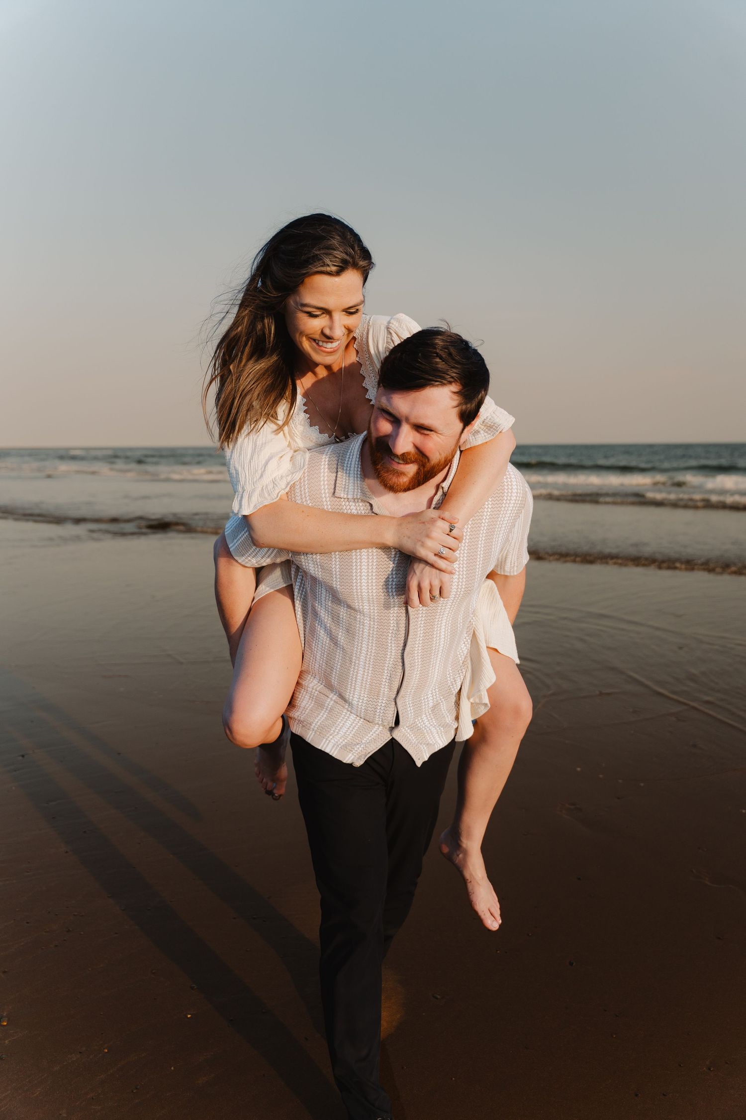 A happy couple enjoys a piggyback ride on a sandy beach at sunset, with ocean waves in the background.