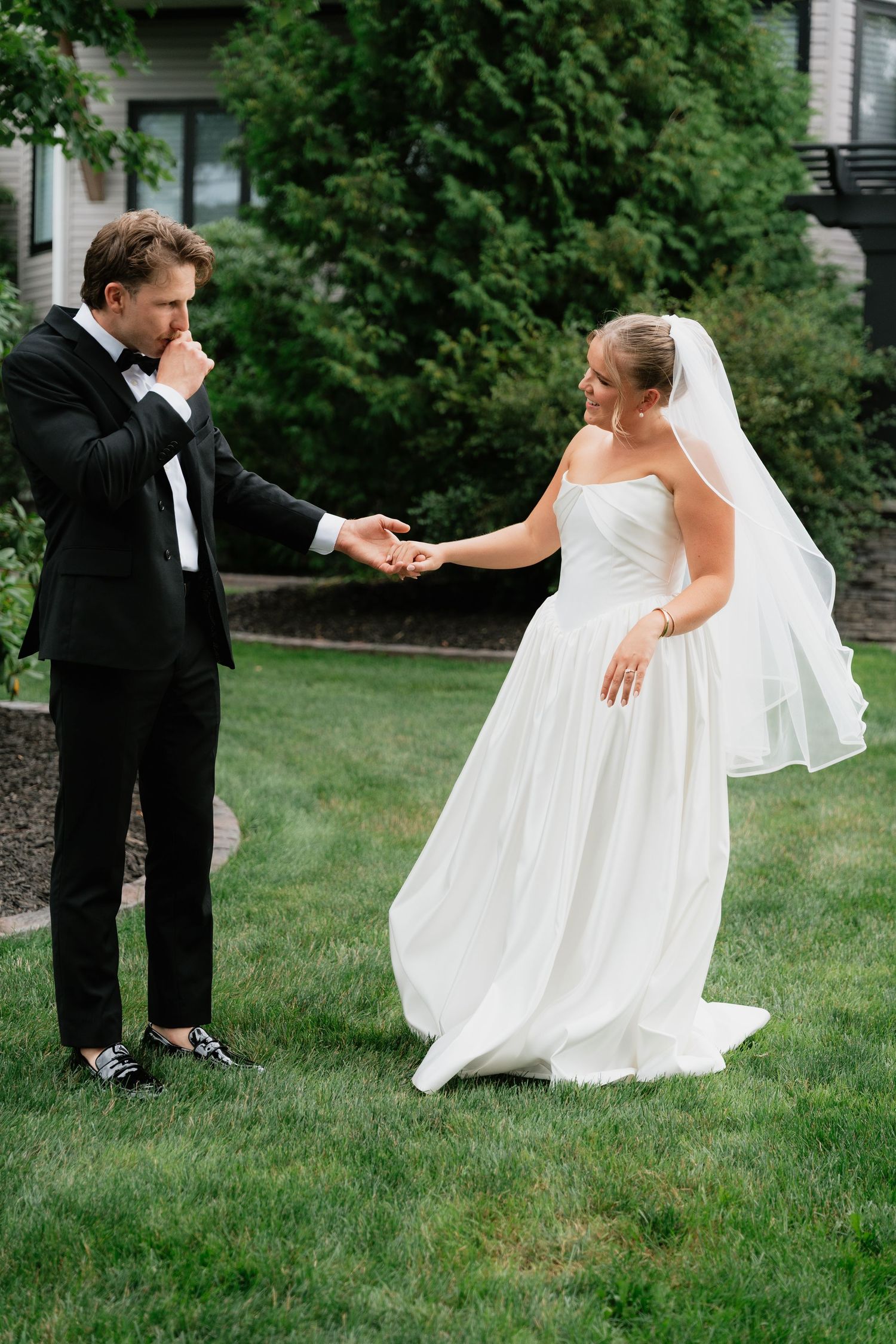 Groom in black tuxedo kisses bride's hand outdoors on green lawn, bride wearing strapless white gown and veil.