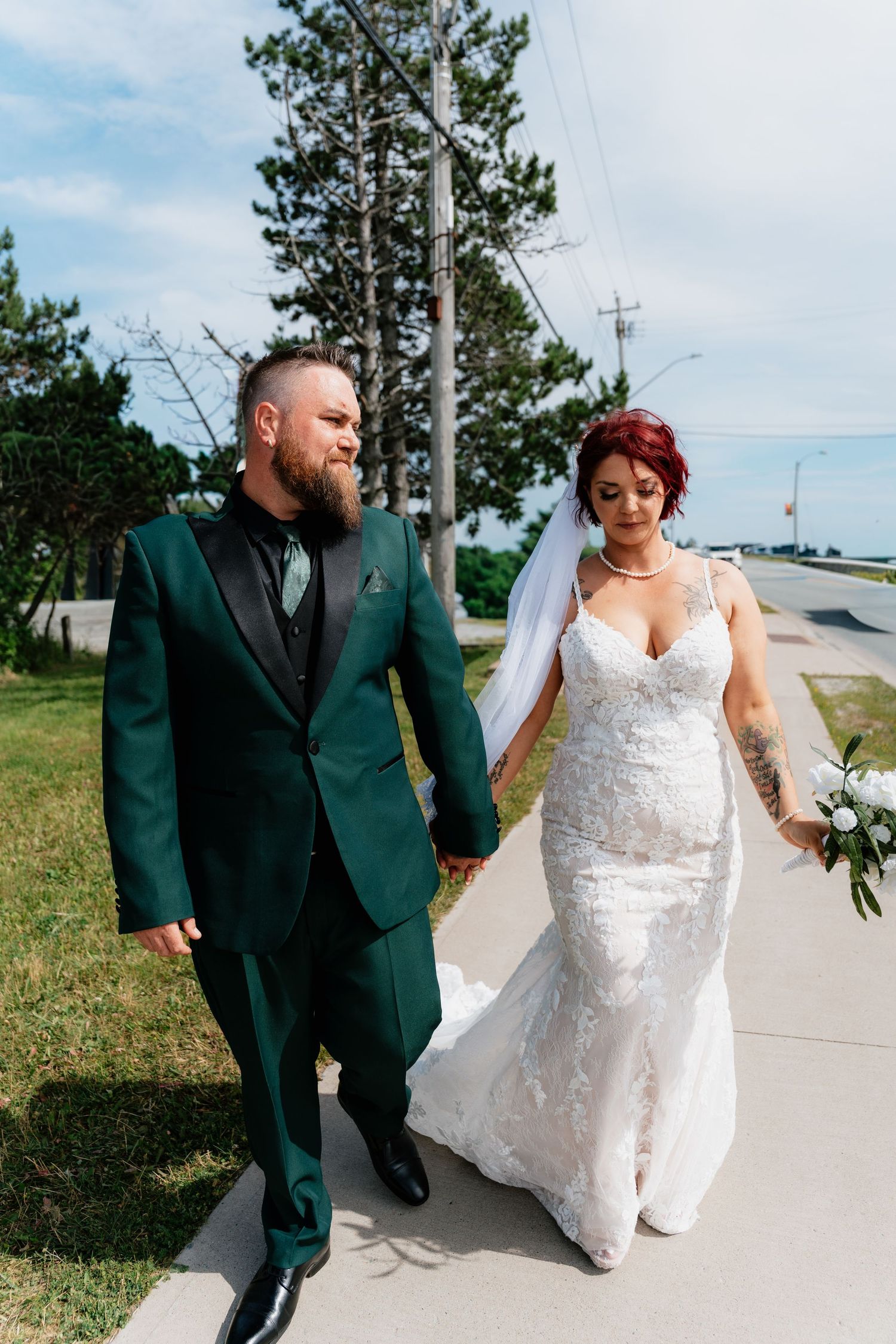 Bride in lace mermaid gown and groom in green tuxedo hold hands walking outdoors on their wedding day.