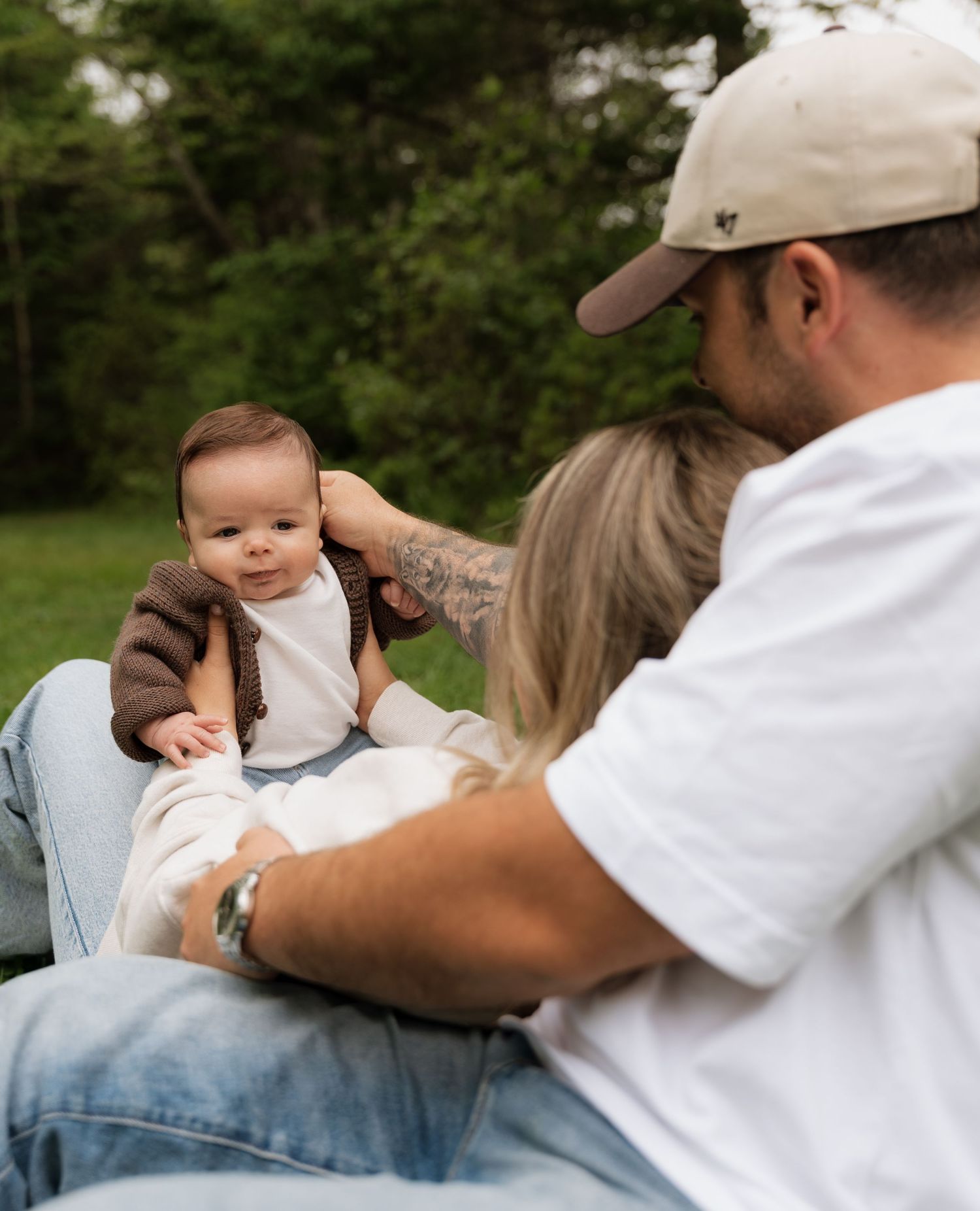 A couple sits on grass holding their smiling baby dressed in a brown cardigan and suspenders outdoors.