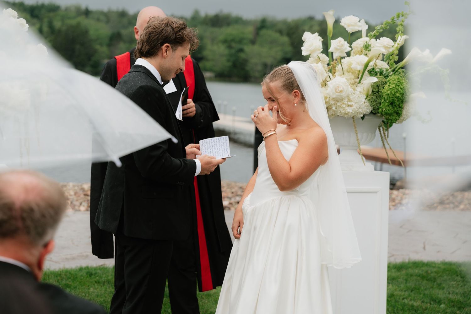 Emotional bride wiping tears during outdoor wedding ceremony with groom reading vows, white floral arrangements, lakeside backdrop.