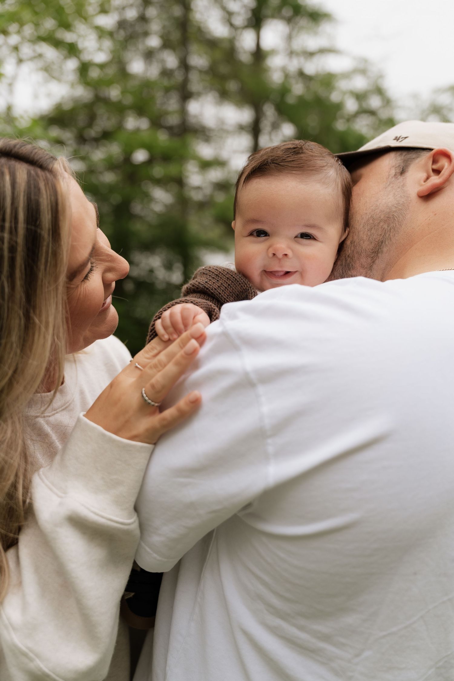 A happy baby smiles while being held by parents outdoors, with lush green trees in the background.