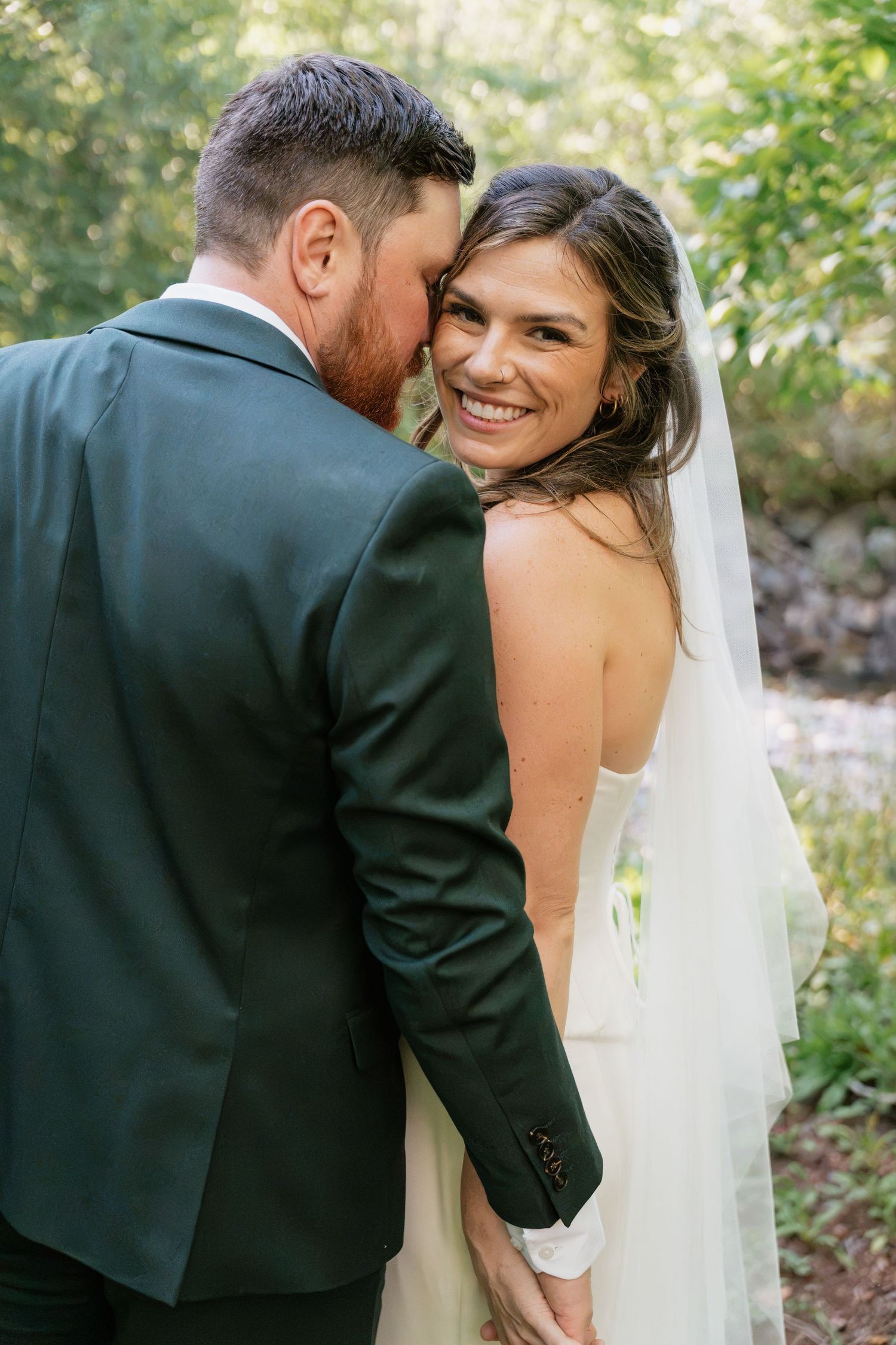 A smiling bride in a white gown and veil embraces her groom in a dark green suit outdoors.