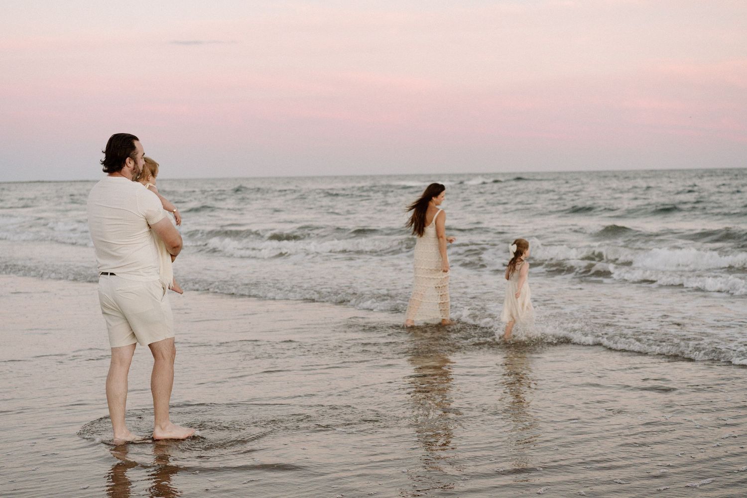 A couple in white outfits walks along a misty beach shoreline during sunset, creating romantic silhouettes against the waves.