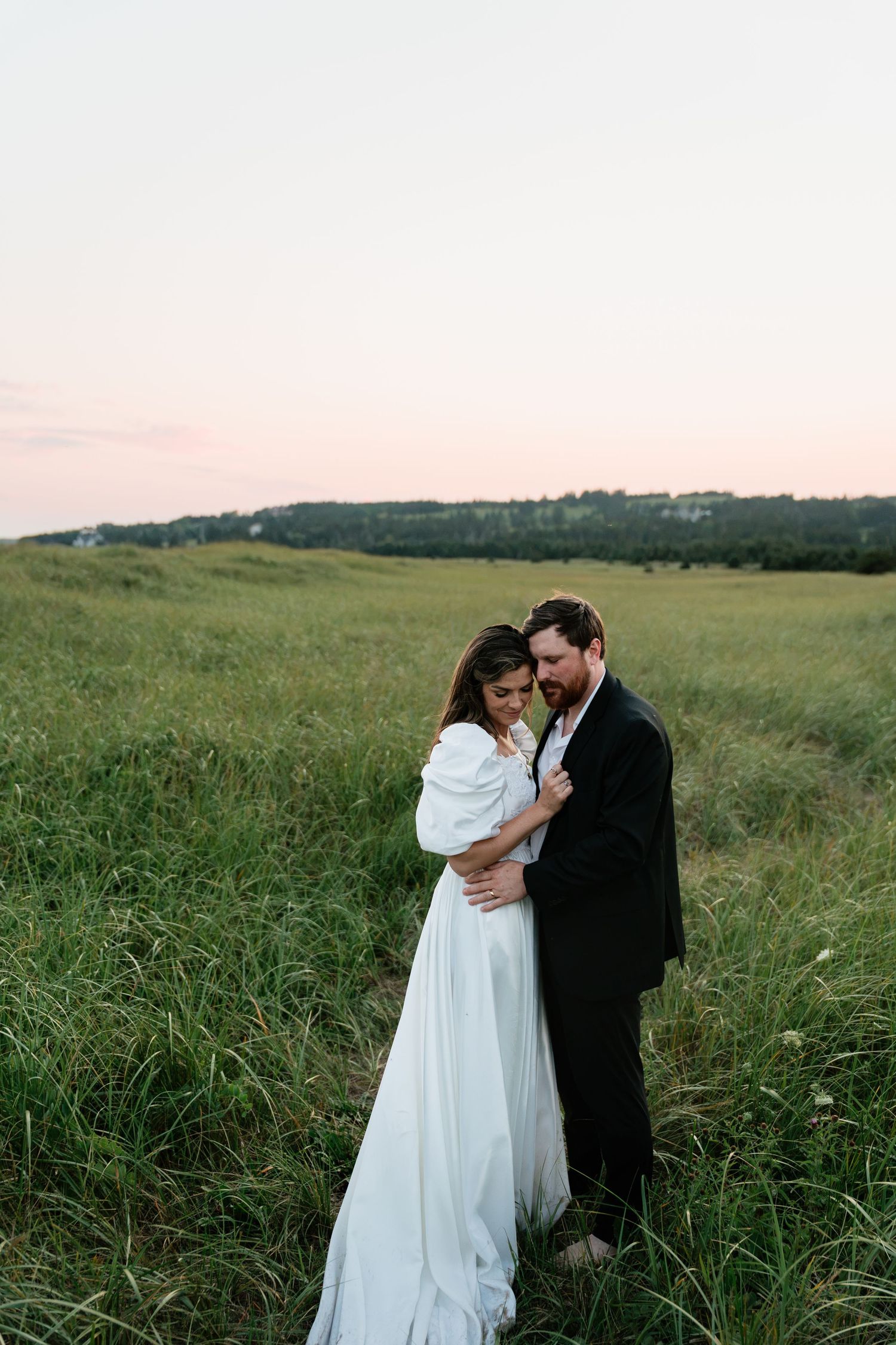 A bride and groom embrace in a lush green field at sunset, she in a white gown, he in a dark suit.