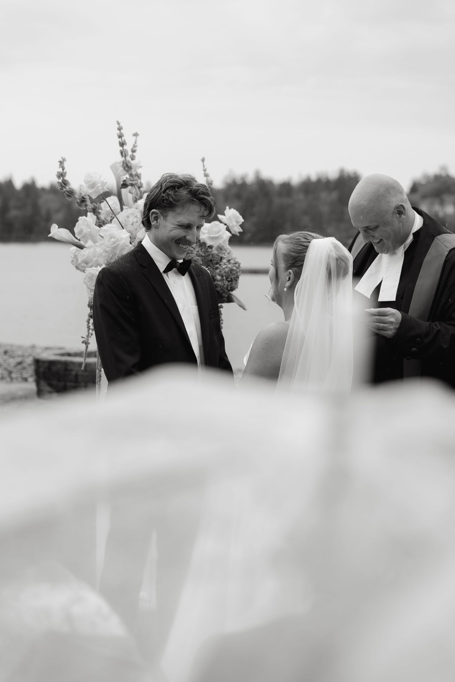 Black and white photo of groom smiling during outdoor lakeside wedding ceremony with officiant.