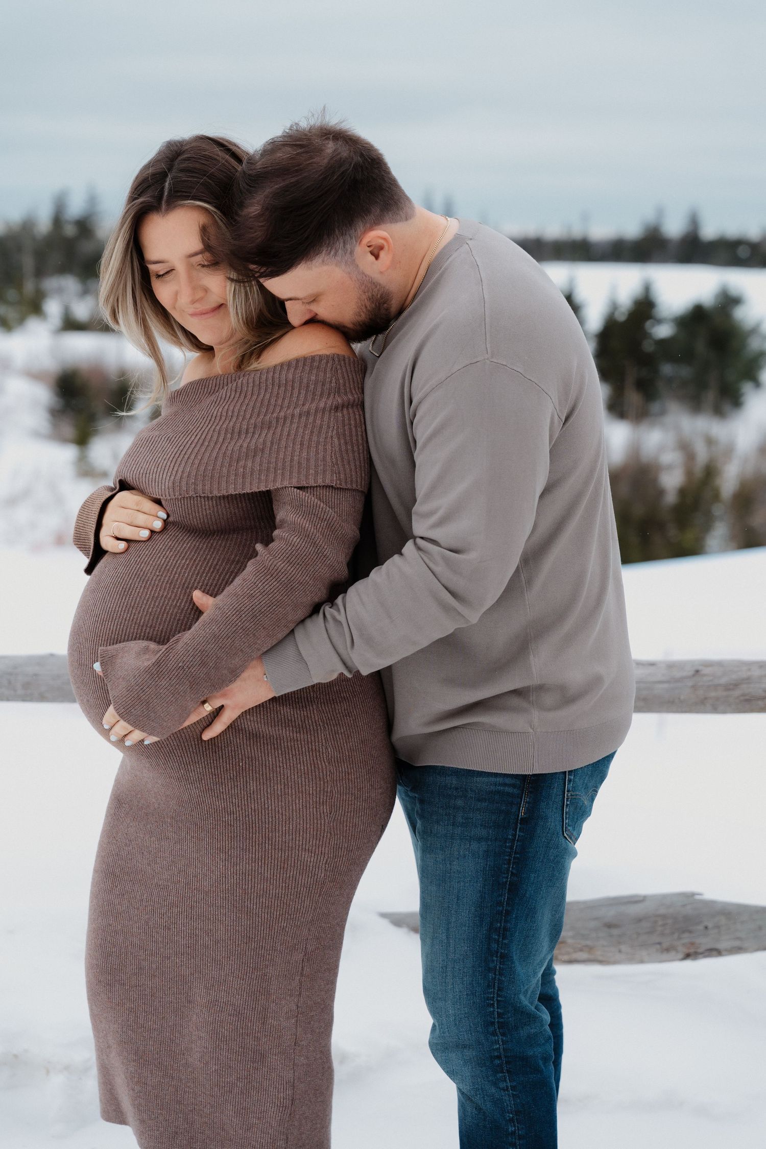 A couple shares an intimate winter maternity photo outdoors in the snow, wearing cozy sweaters.
