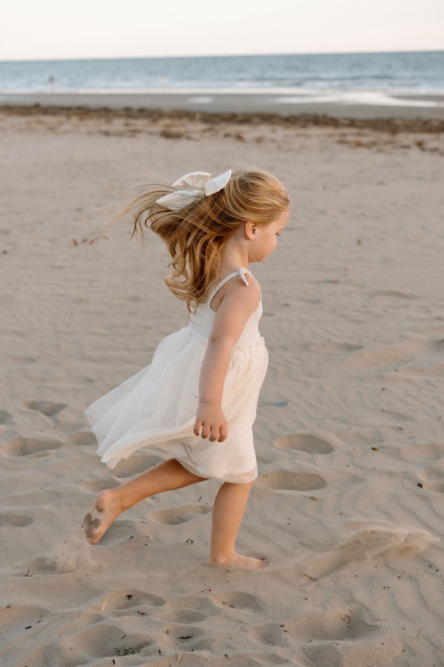Young child in a flowing white dress runs playfully on a sandy beach.