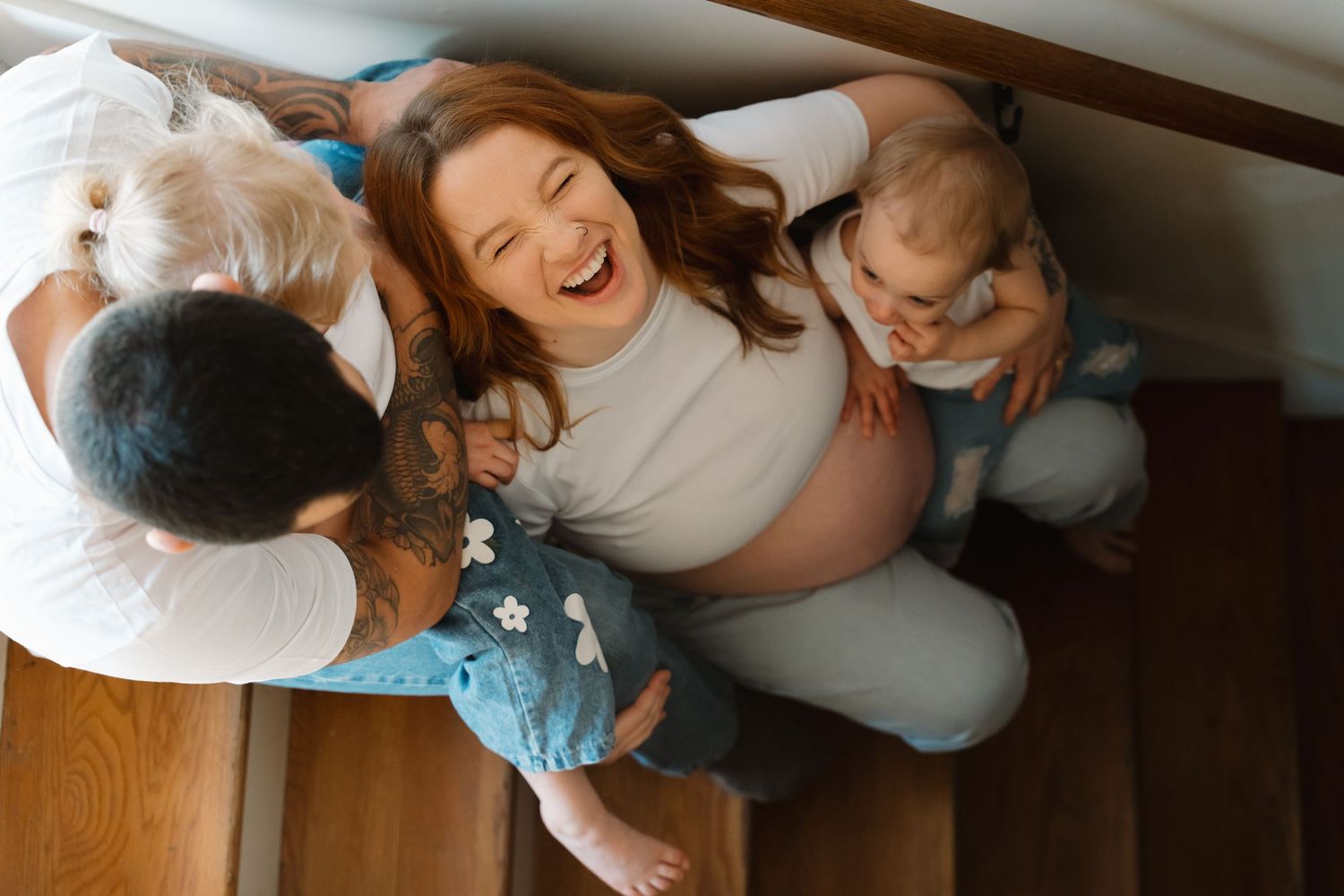 Happy pregnant woman laughing with family on stairs, holding baby while partner looks on in warm home setting.