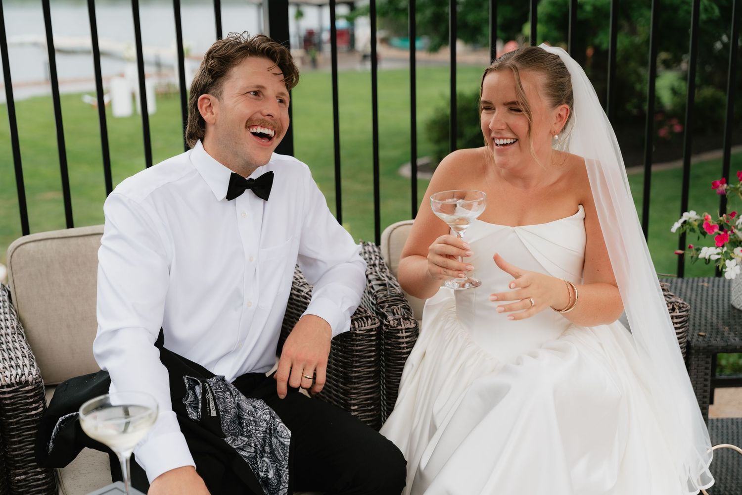 Laughing bride in white gown and groom in tuxedo sitting together outdoors at their wedding reception.