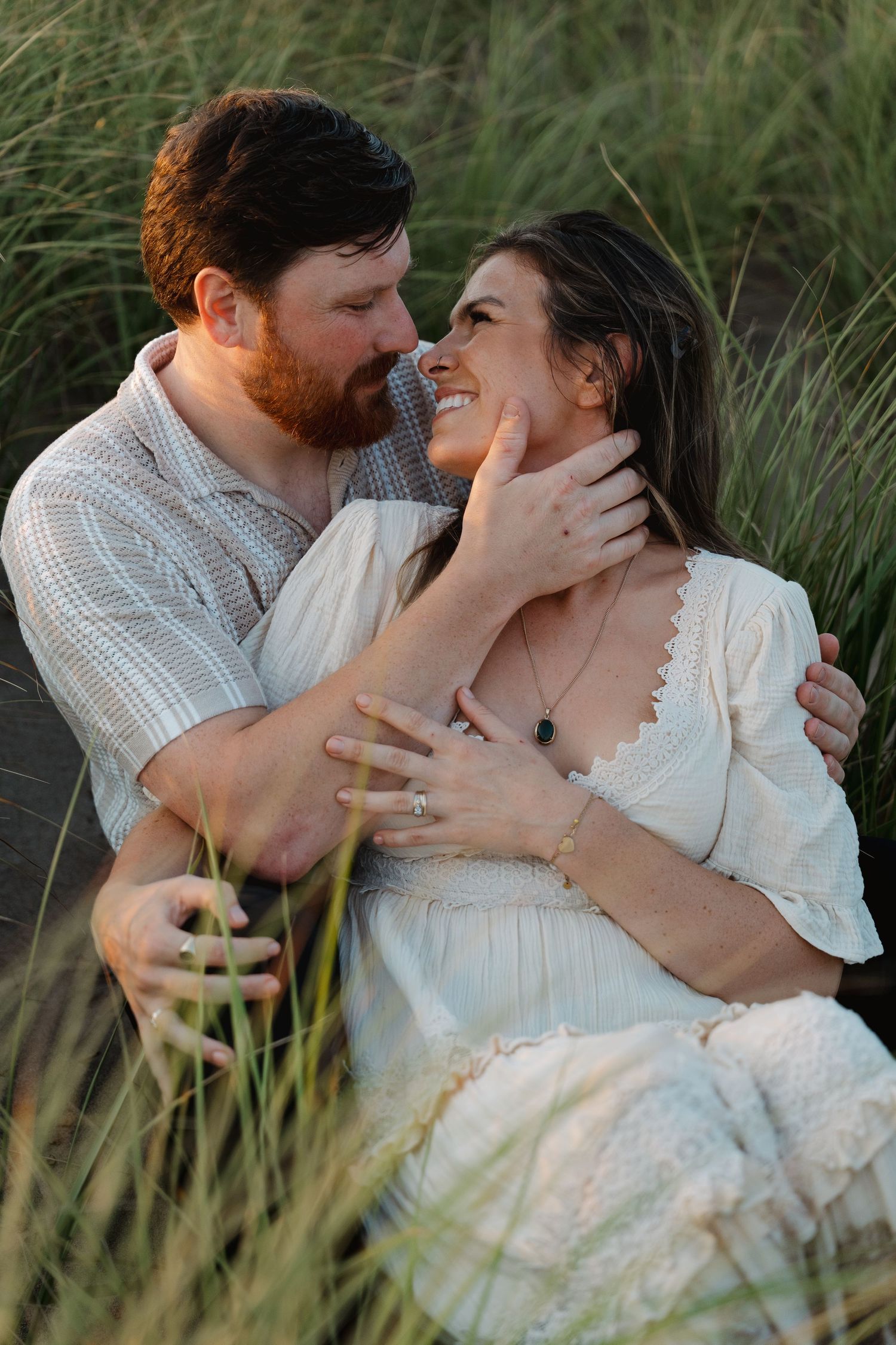 Couple sitting in tall grass, smiling and gazing at each other during a romantic outdoor engagement photo session.