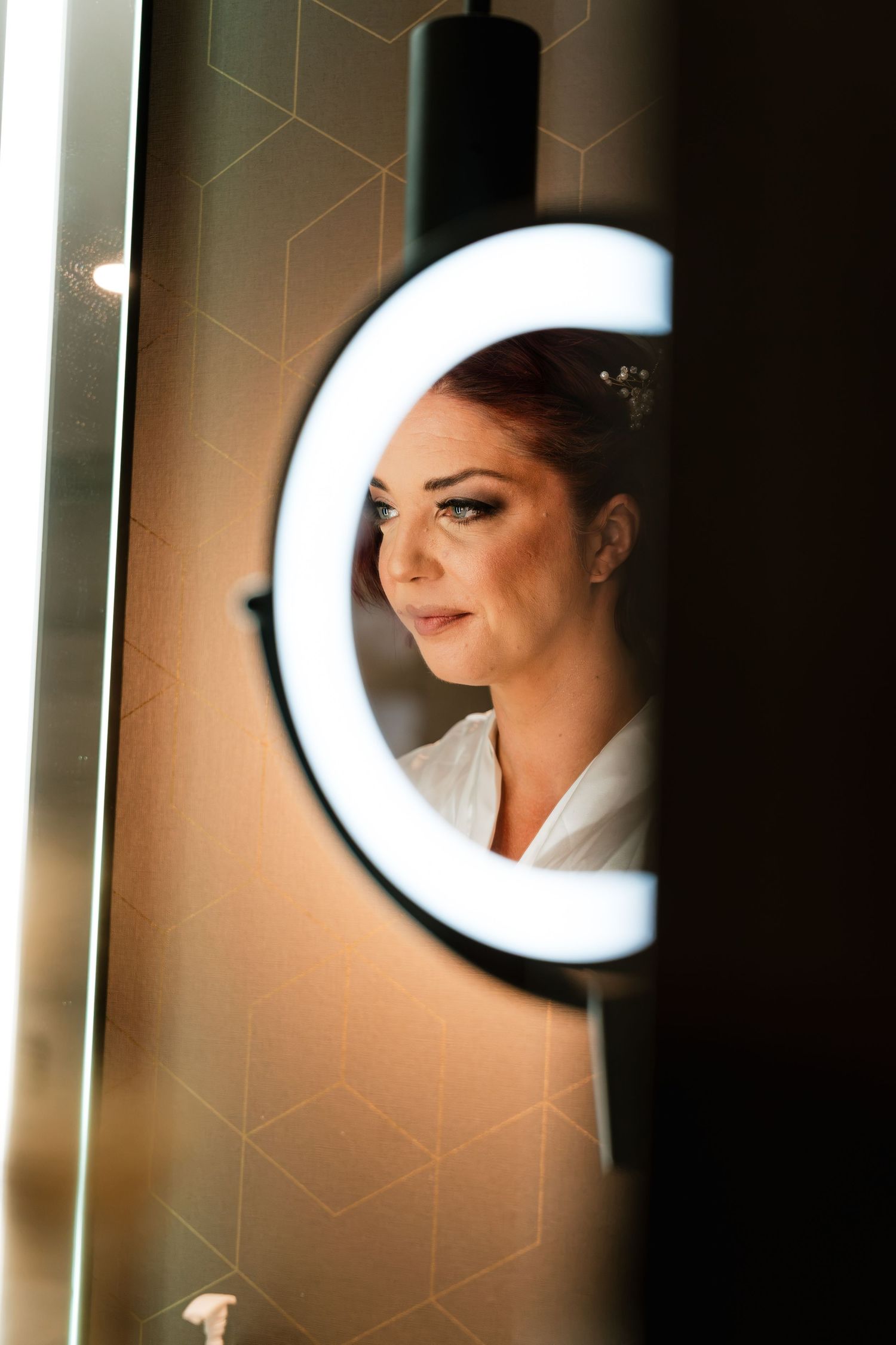 Bride with red hair and elegant makeup reflected in a round illuminated vanity mirror while getting ready.