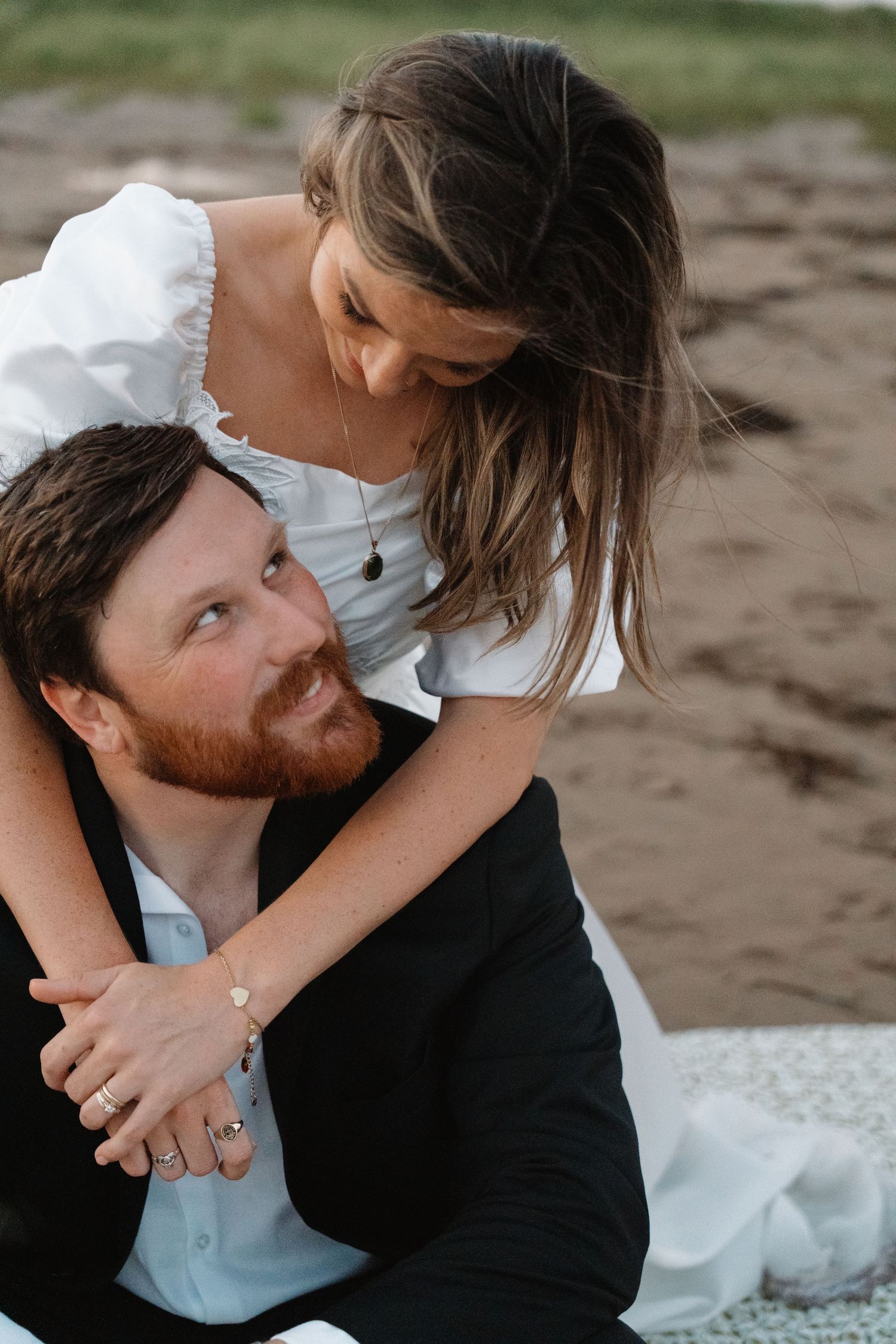A couple shares a tender moment on a sandy beach, woman in white dress embracing red-bearded man from behind.