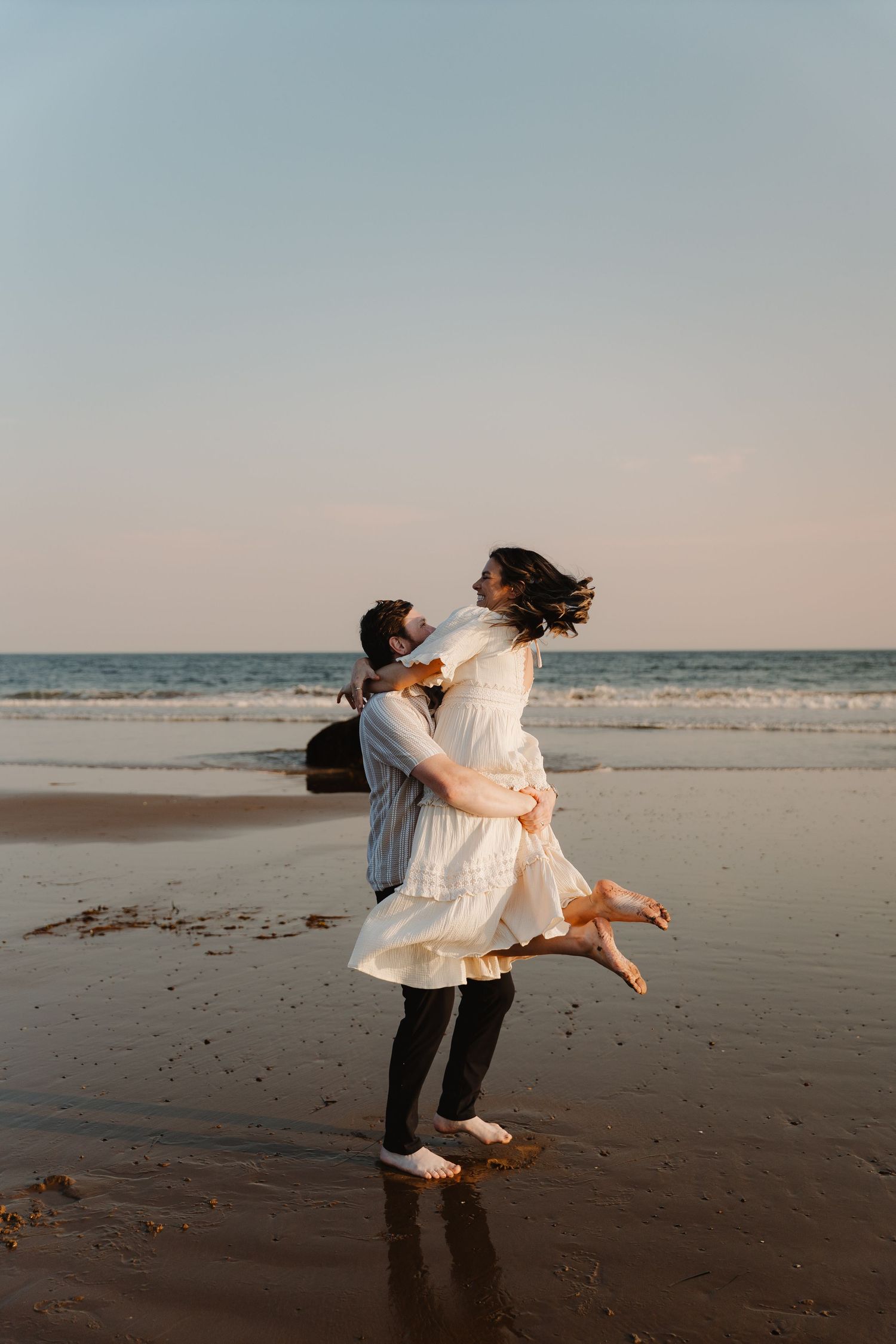 Couple sharing a romantic moment on the beach, man lifting woman in white dress at sunset by the ocean.