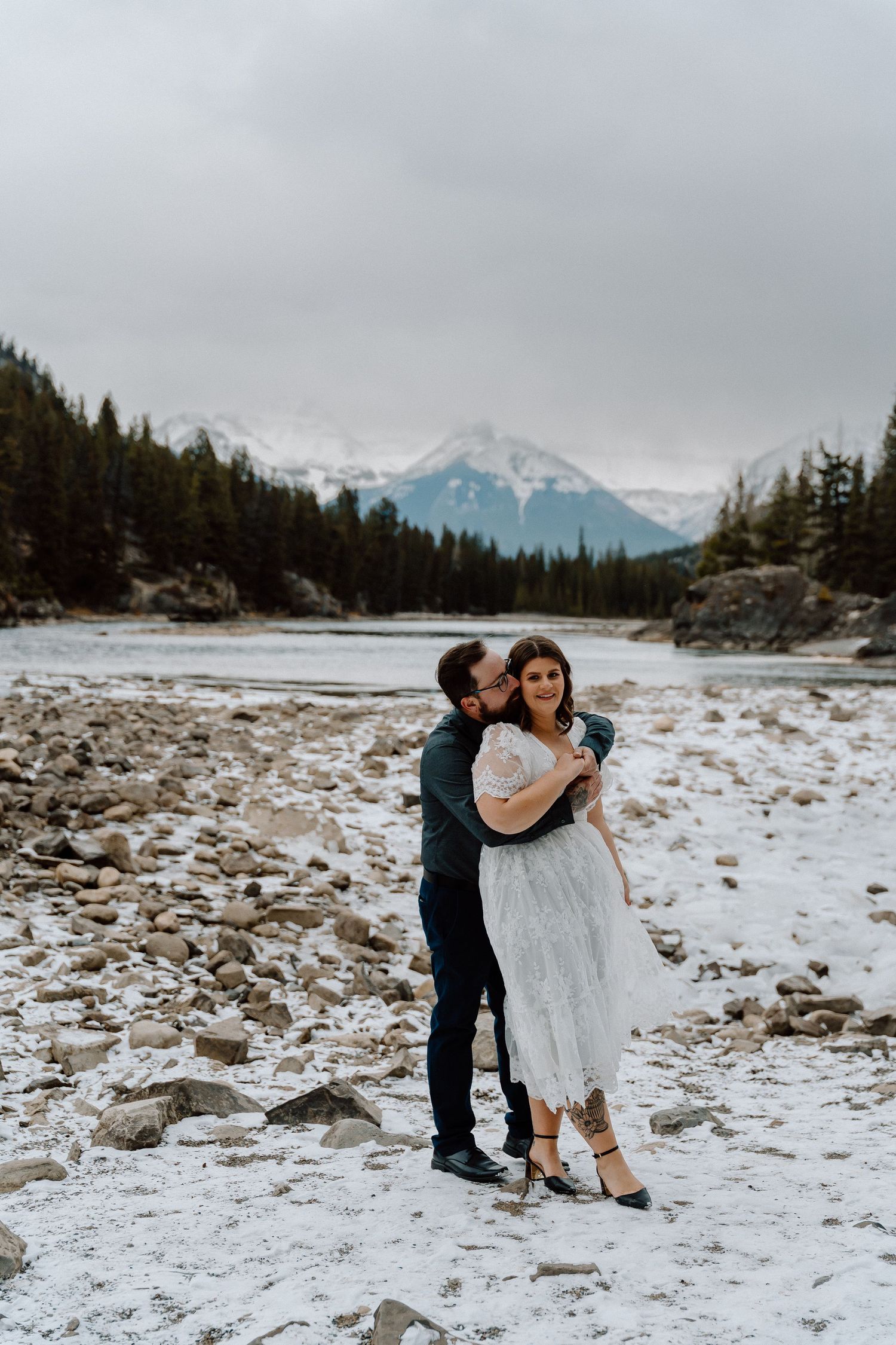 Winter wedding photos on rocky snow-covered shore with mountain peaks in distance.