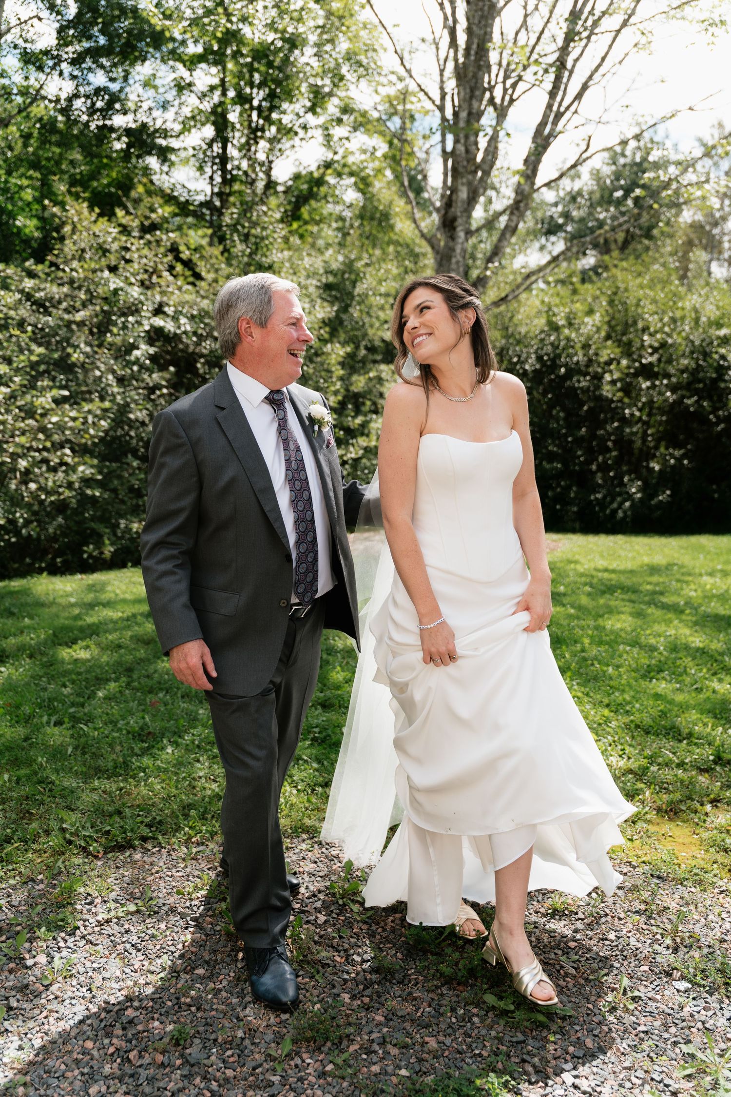 A bride in a white gown walks with an older man in a gray suit through a lush garden, both smiling joyfully.