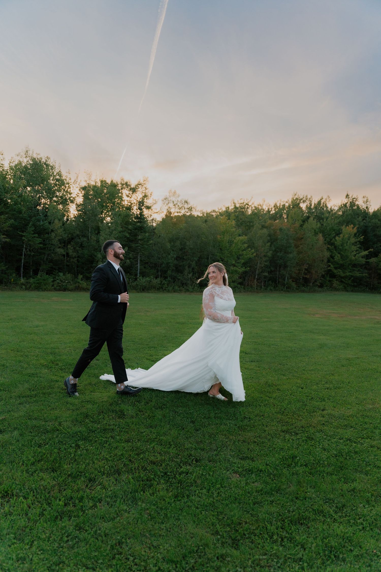 A bride and groom walk hand in hand across a lush green field at sunset, surrounded by trees.