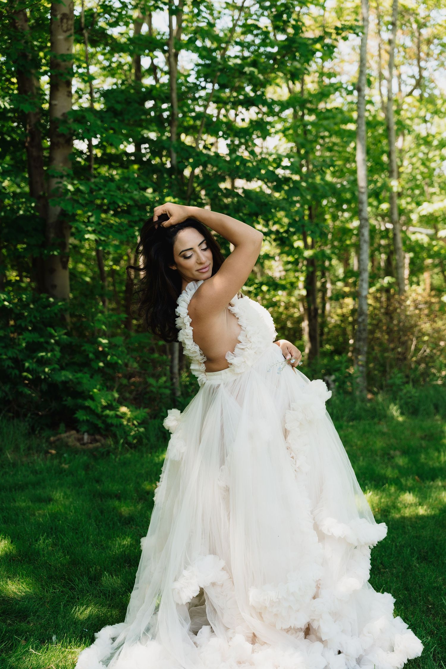 A bride in a flowing white tulle gown with floral details poses gracefully outdoors in a lush green forest setting.