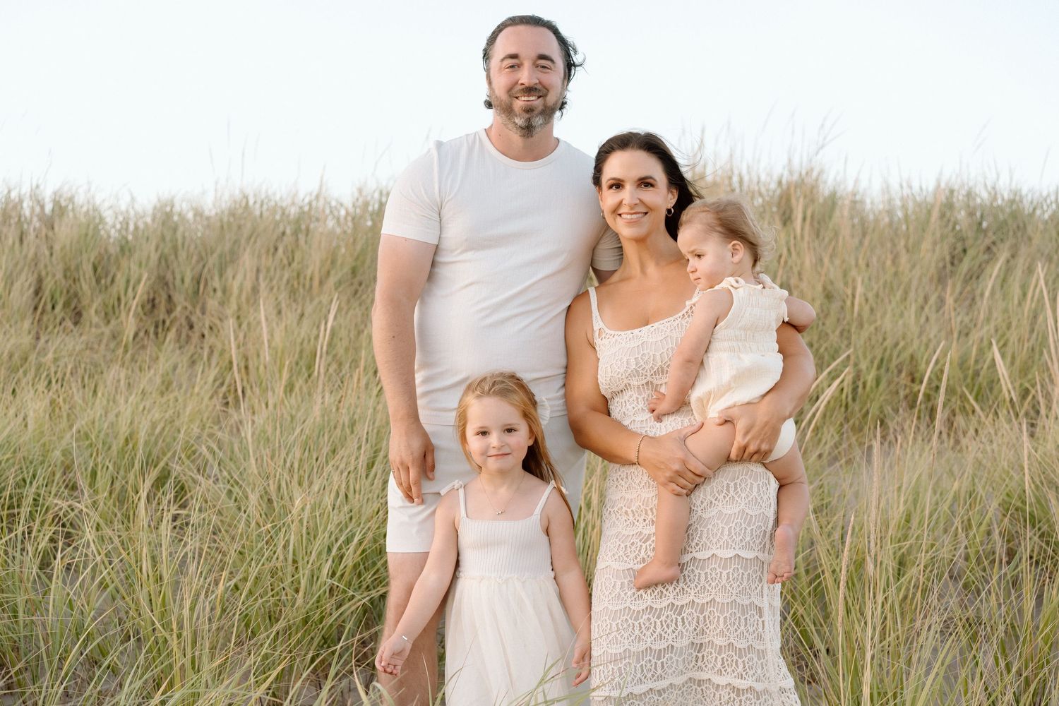 Family dressed in white and neutral colors poses together on grassy coastal dunes.