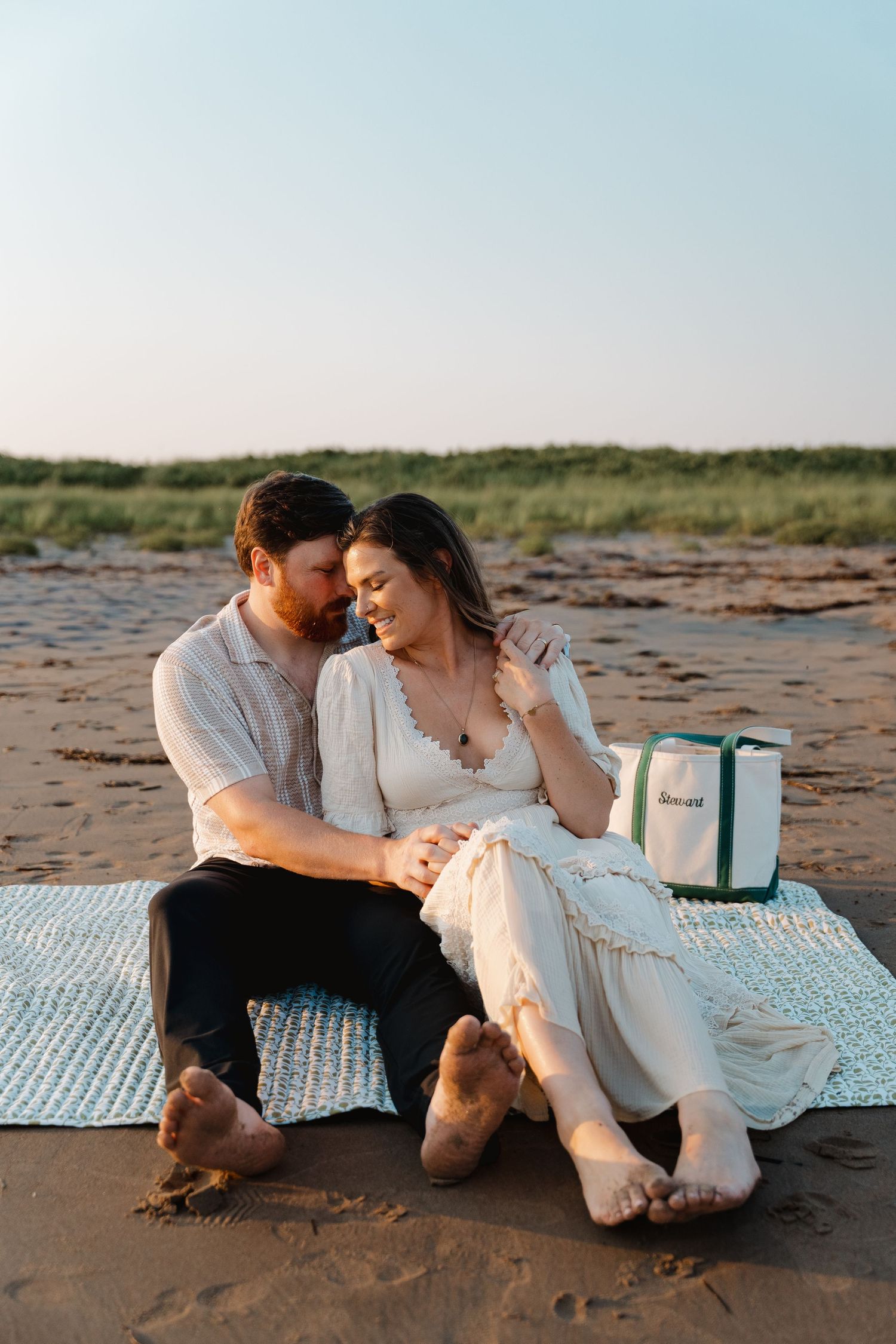 Couple sitting barefoot on a blanket at the beach during golden hour, sharing a tender romantic moment.