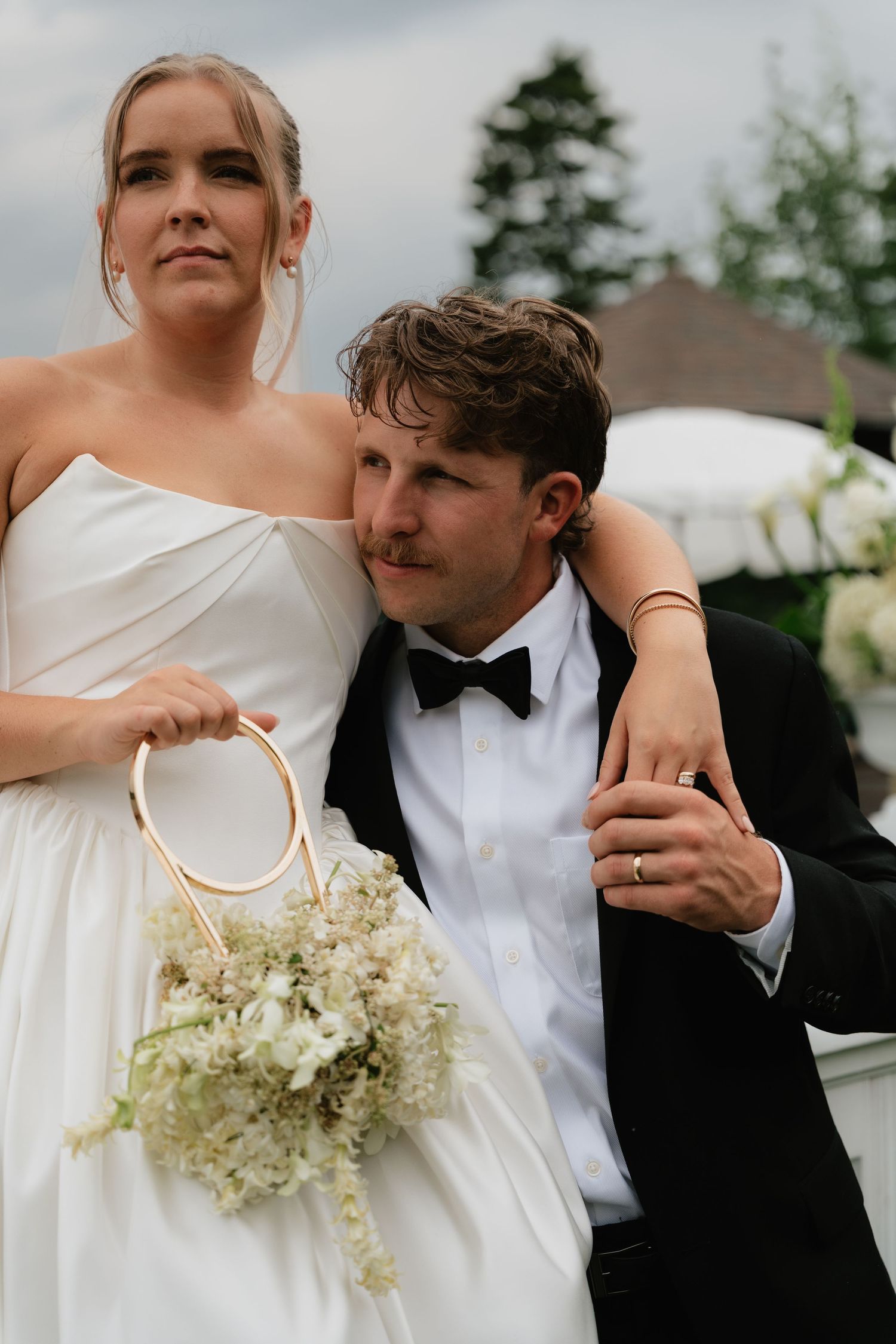 Bride in white gown holding floral bouquet stands beside groom in black tuxedo at outdoor wedding ceremony.