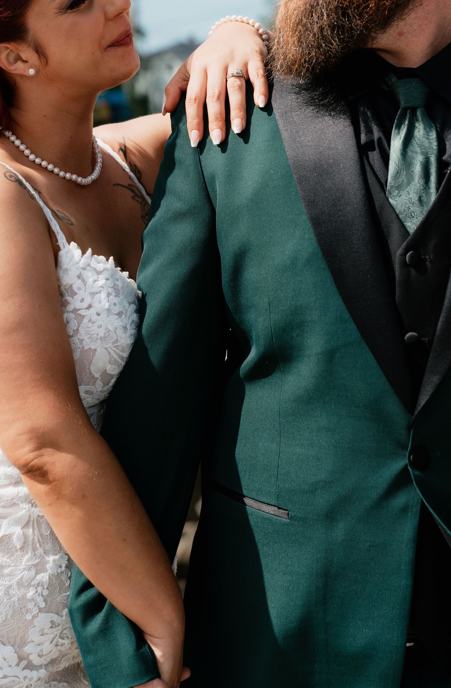 Bride in white lace gown rests hand with ring on groom's green tuxedo jacket with black lapels at wedding.