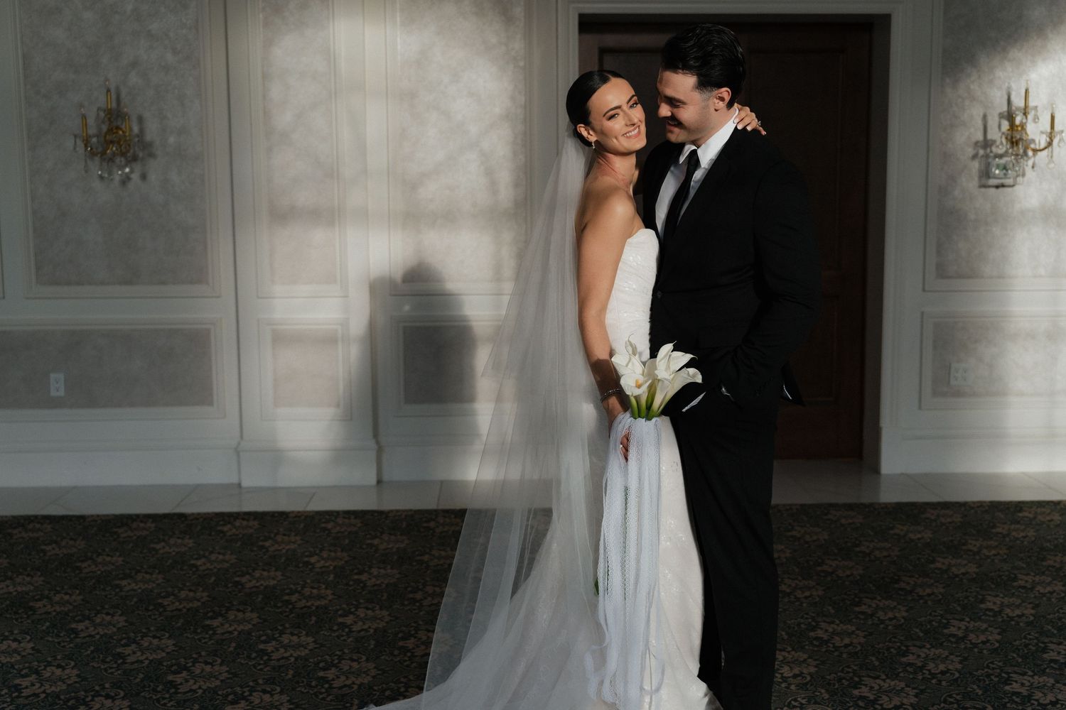 A romantic couple shares an intimate moment in an elegant ballroom setting with ornate chandeliers and white decor.
