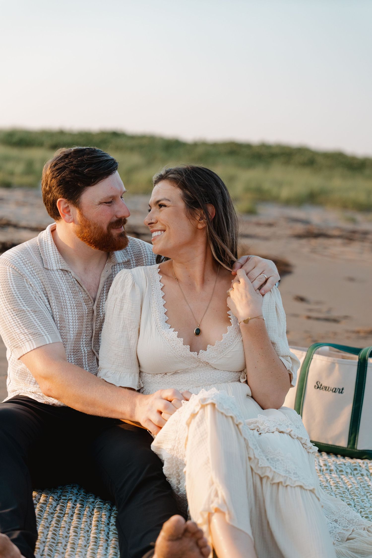 A couple sits together on a beach blanket at sunset, smiling warmly at each other near a monogrammed bag.