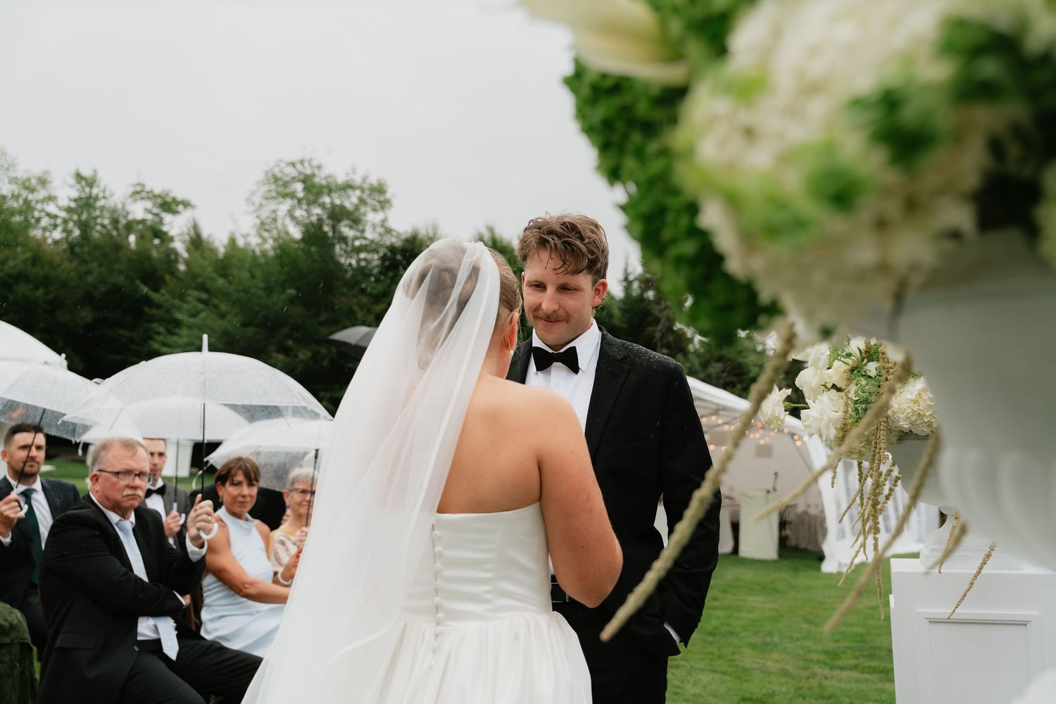 Bride and groom exchange vows at outdoor wedding ceremony with white floral arrangements and guests holding umbrellas.