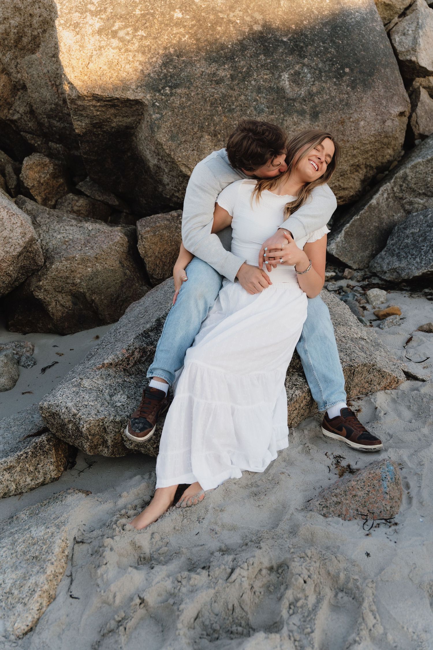Couple sitting on beach rocks, man in white shirt embracing laughing woman in white dress and jeans.