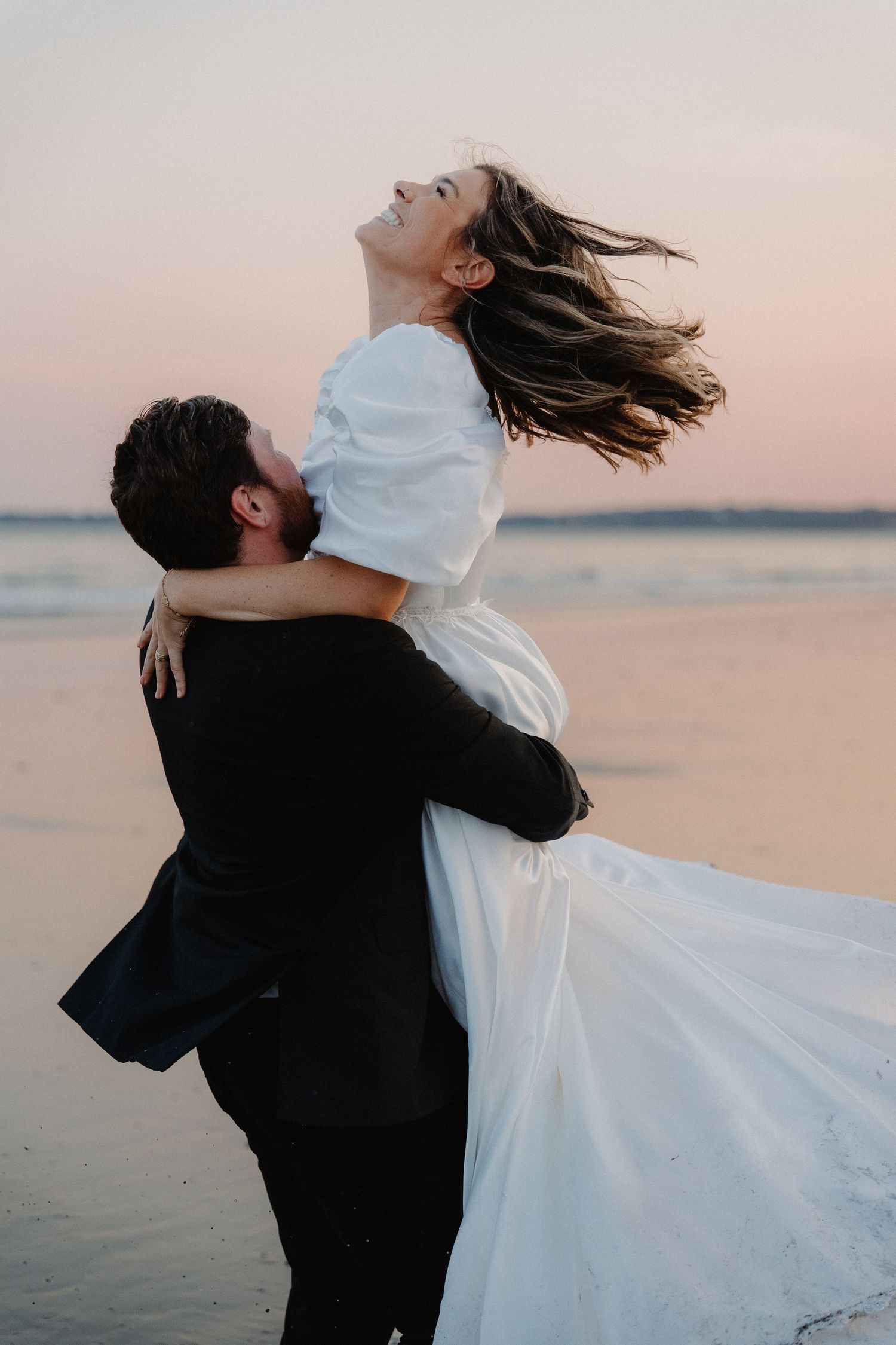 Groom lifting bride in white dress on beach at sunset, her hair flowing as she laughs joyfully.