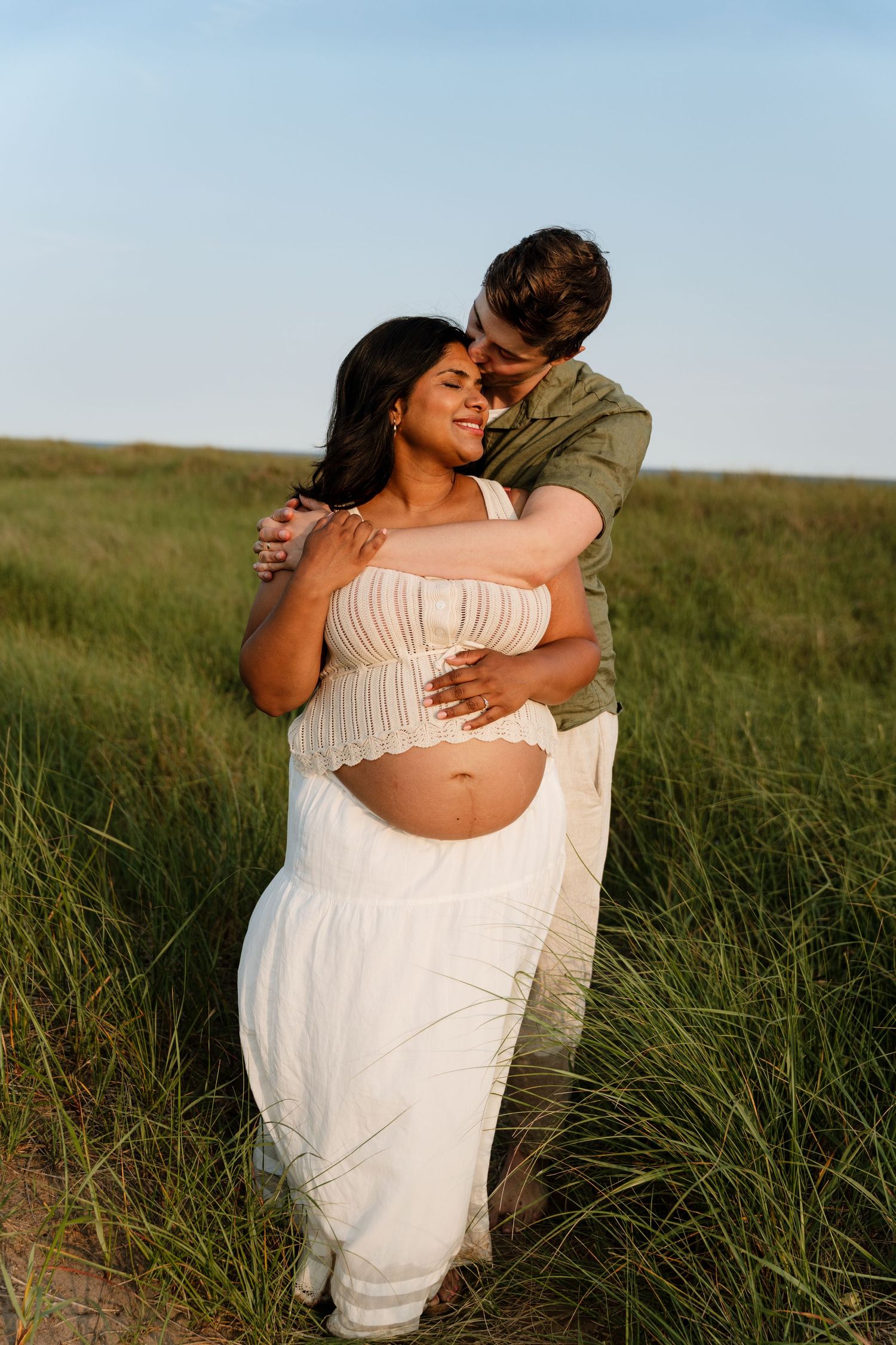 Couple embracing in green grass field, pregnant woman in white skirt and crop top, partner in green shirt, golden hour.