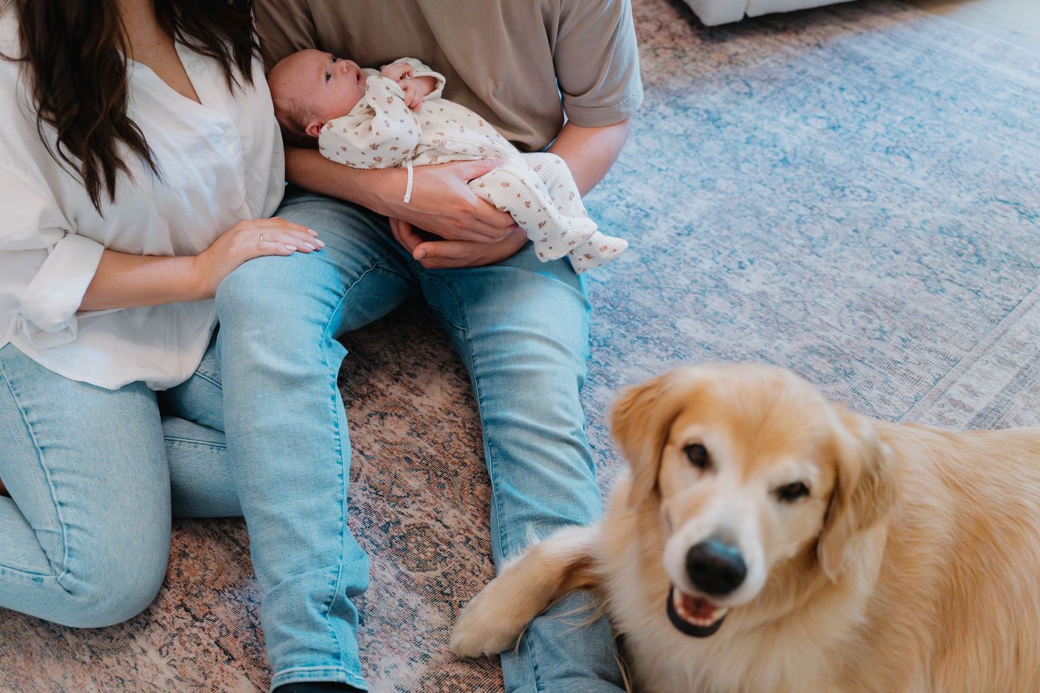 A couple sits on the floor holding a newborn baby while a golden retriever dog lies beside them indoors.