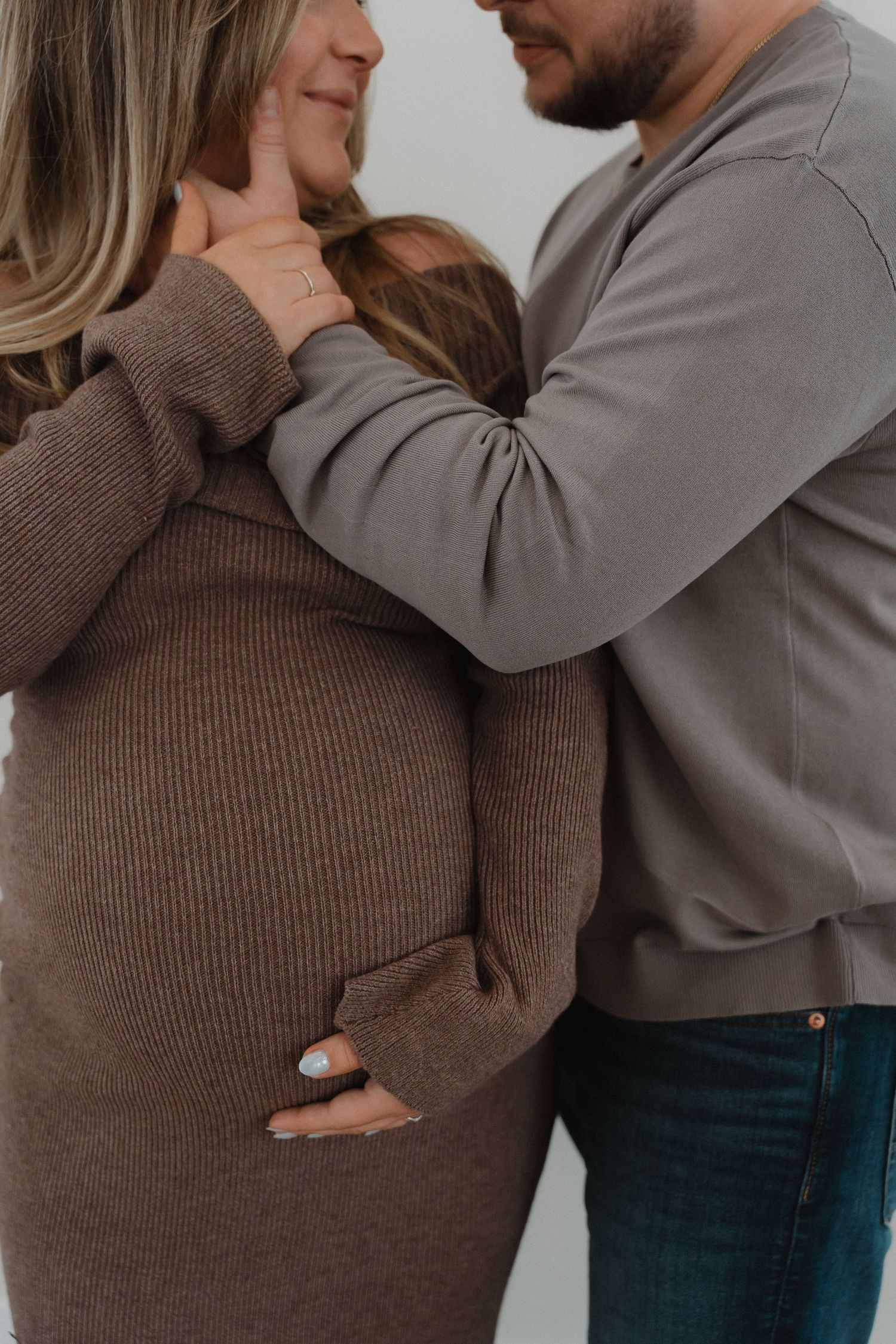 Intimate maternity photo showing couple in cozy sweaters embracing with hands on pregnant belly against white wall.