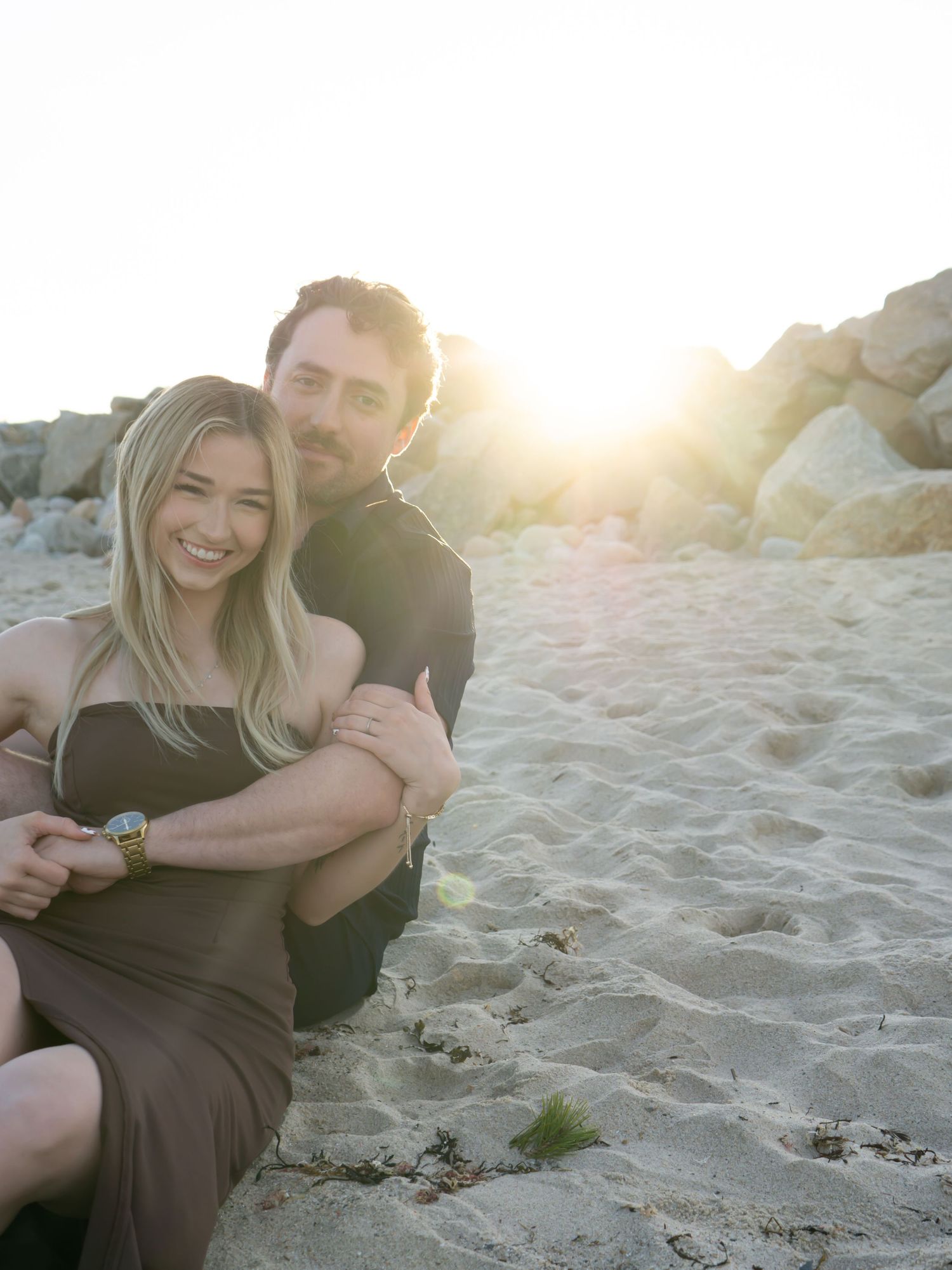Romantic beach engagement photos at sunset with couple sitting in golden sand.