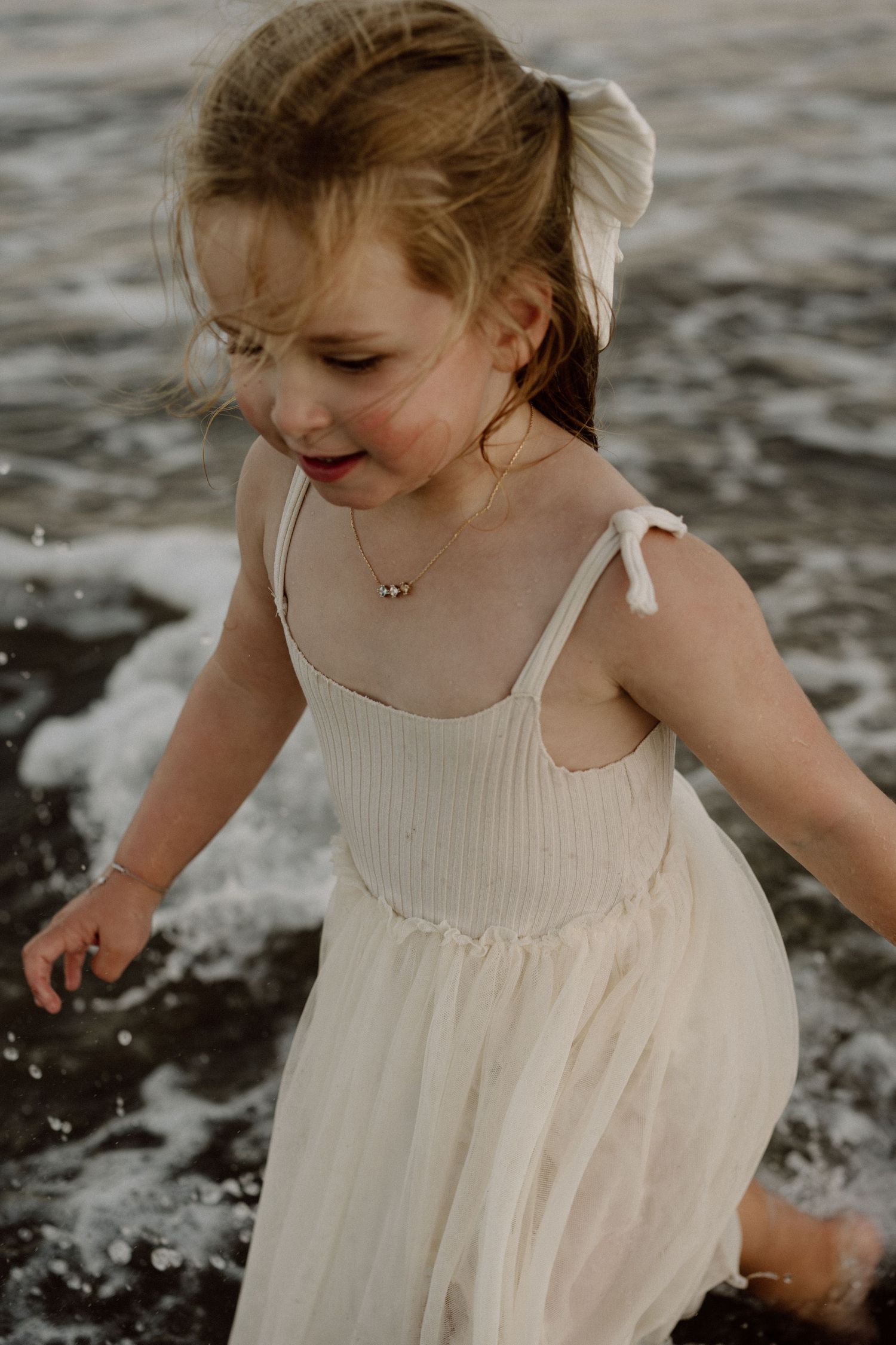 Child in white sundress plays joyfully by the ocean waves.