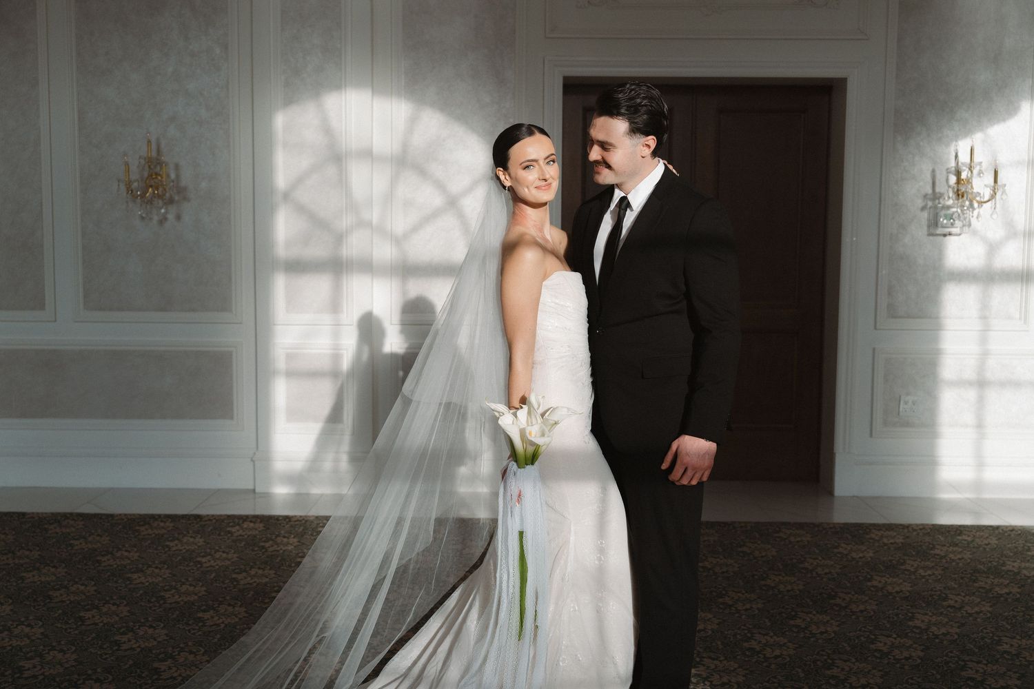 A bride and groom share an intimate moment in an elegant white ballroom with arched windows and ornate details.