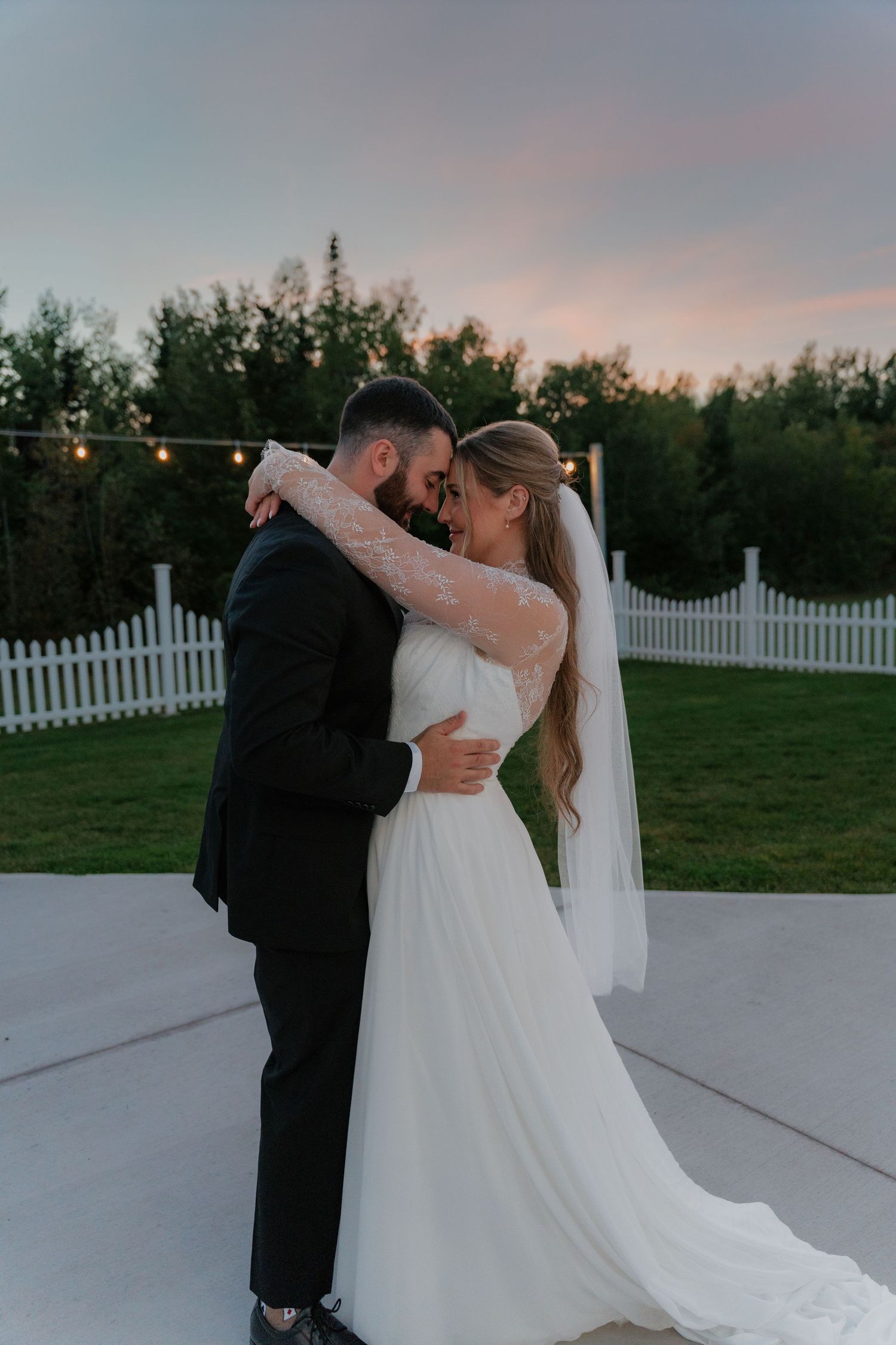 A bride and groom share a romantic embrace at sunset outdoors, with white picket fence and trees in the background.