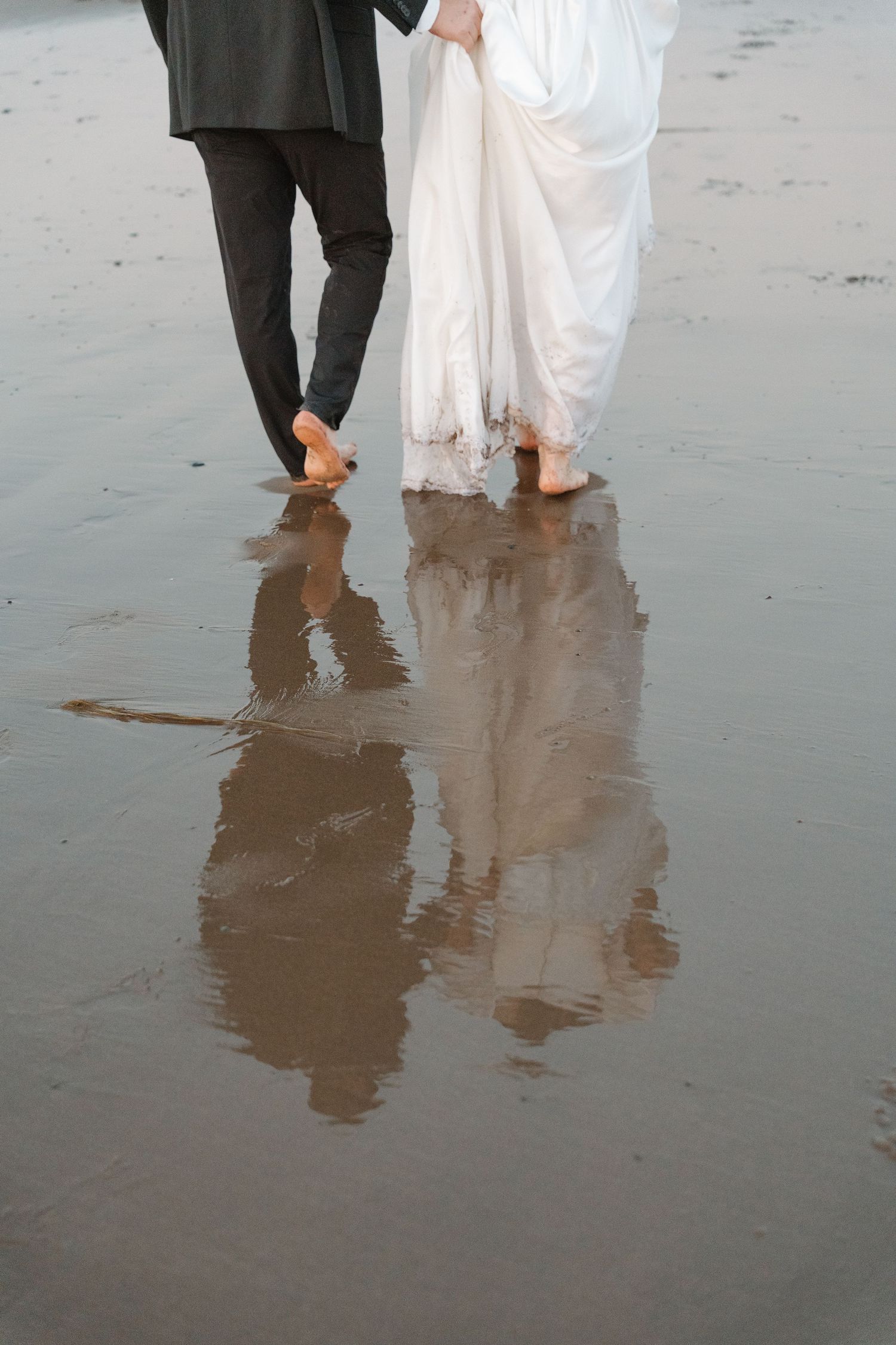 Barefoot bride and groom walking on wet sandy beach, their reflections visible in the water below.
