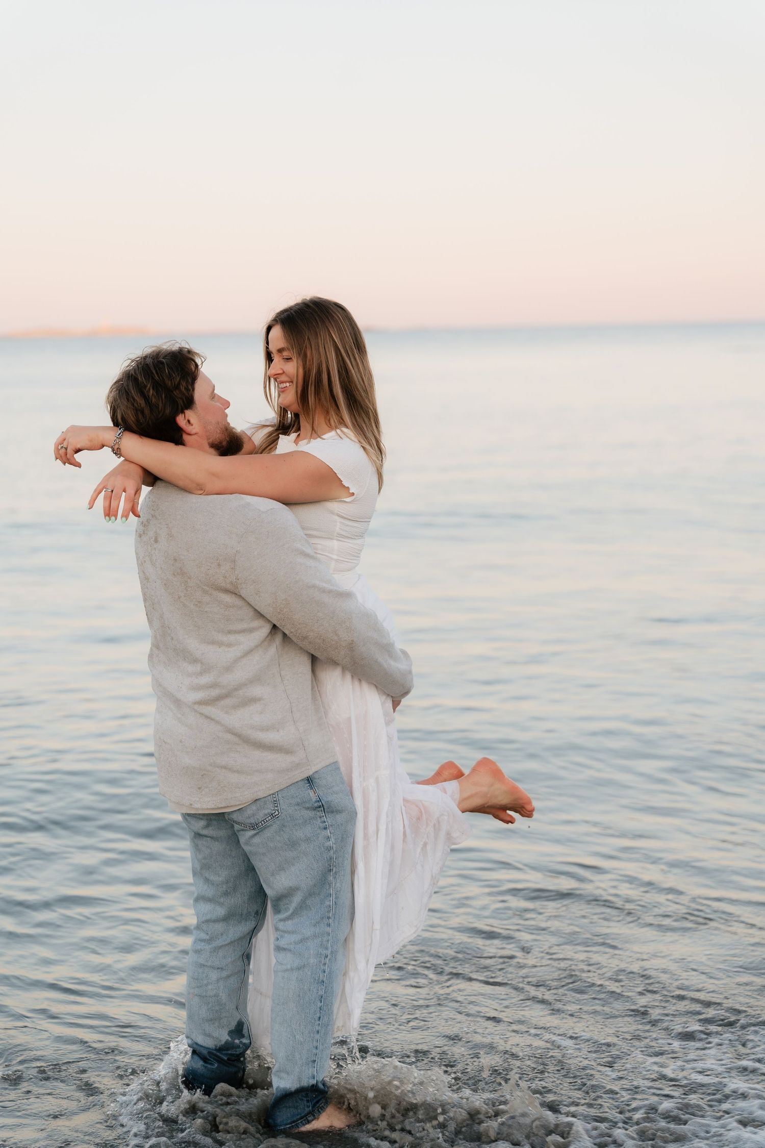 A couple embraces romantically at the beach during sunset, man lifting woman in white dress while waves lap at their feet.