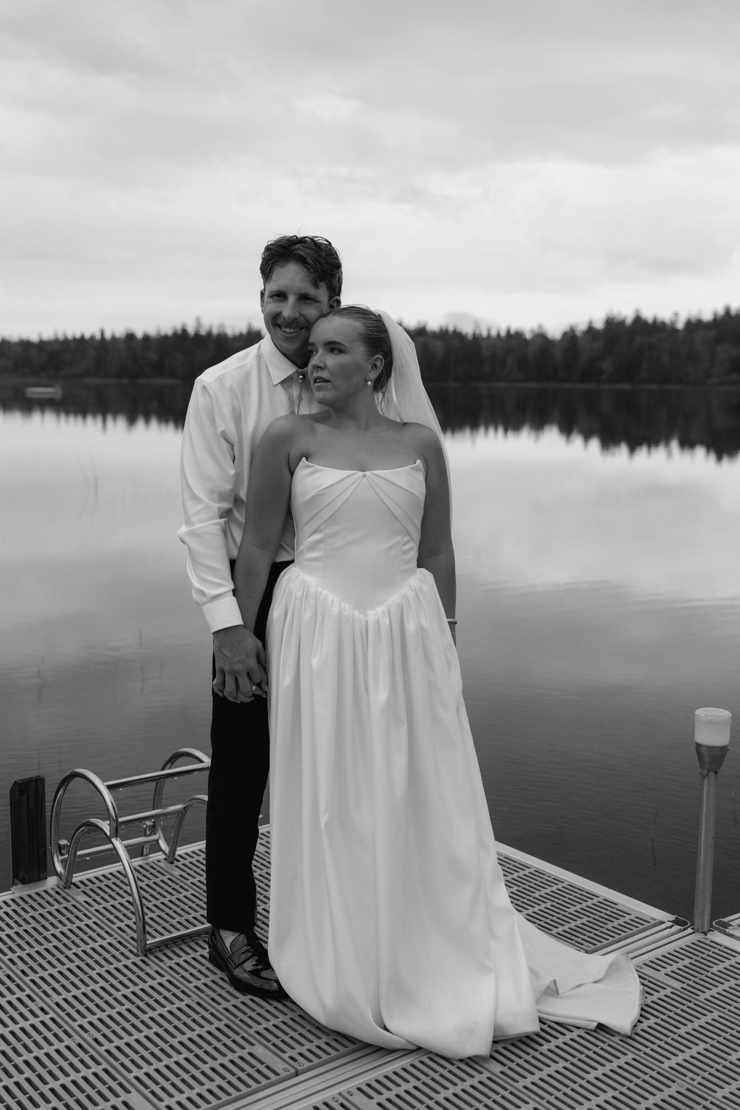 Black and white portrait of bride and groom holding hands on a dock with lake backdrop.