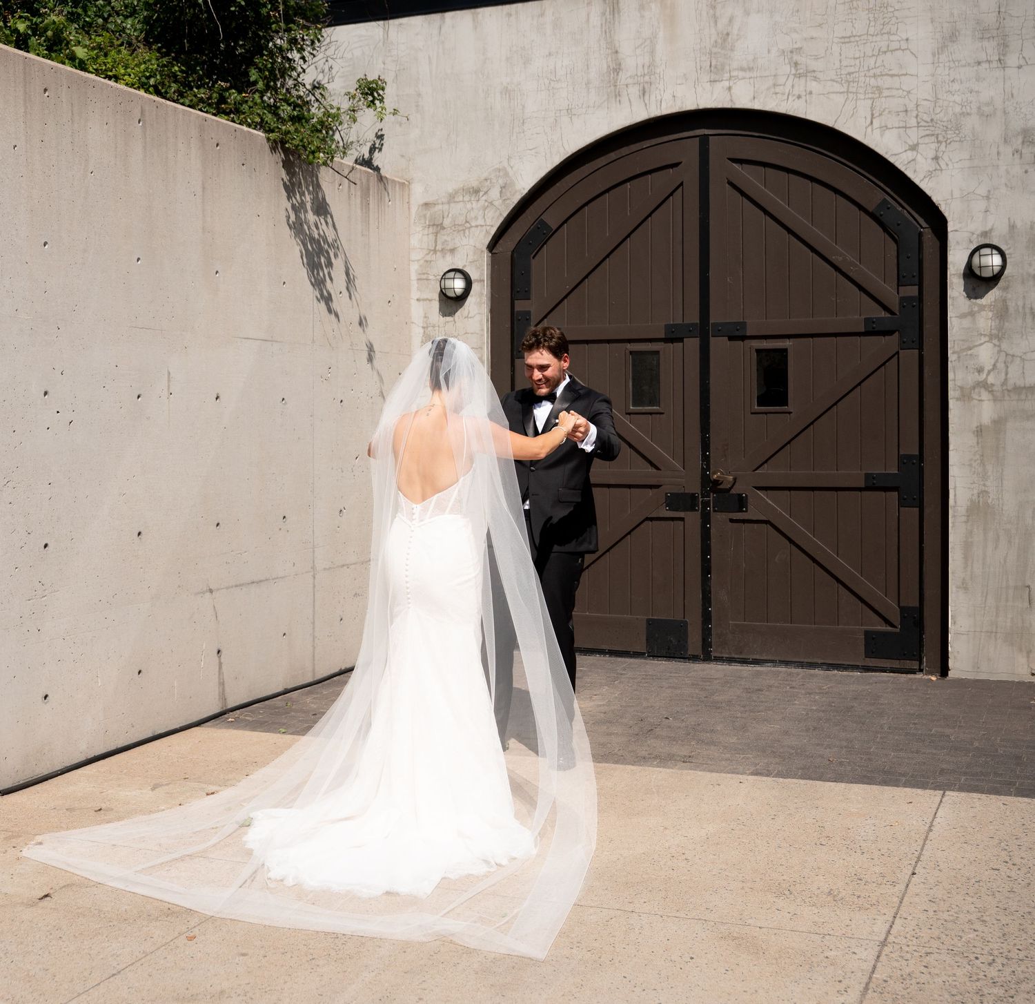 A bride in a white gown with long veil and groom in black tuxedo share a first look moment before large wooden doors.