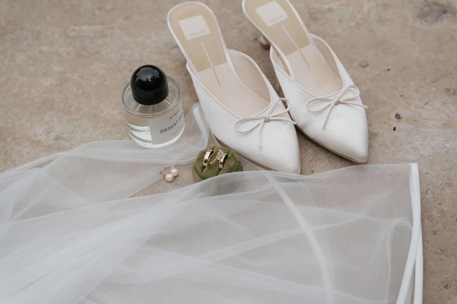 Elegant bridal flat shoes with bow, perfume bottle, gold rings, and sheer veil arranged on a neutral surface.
