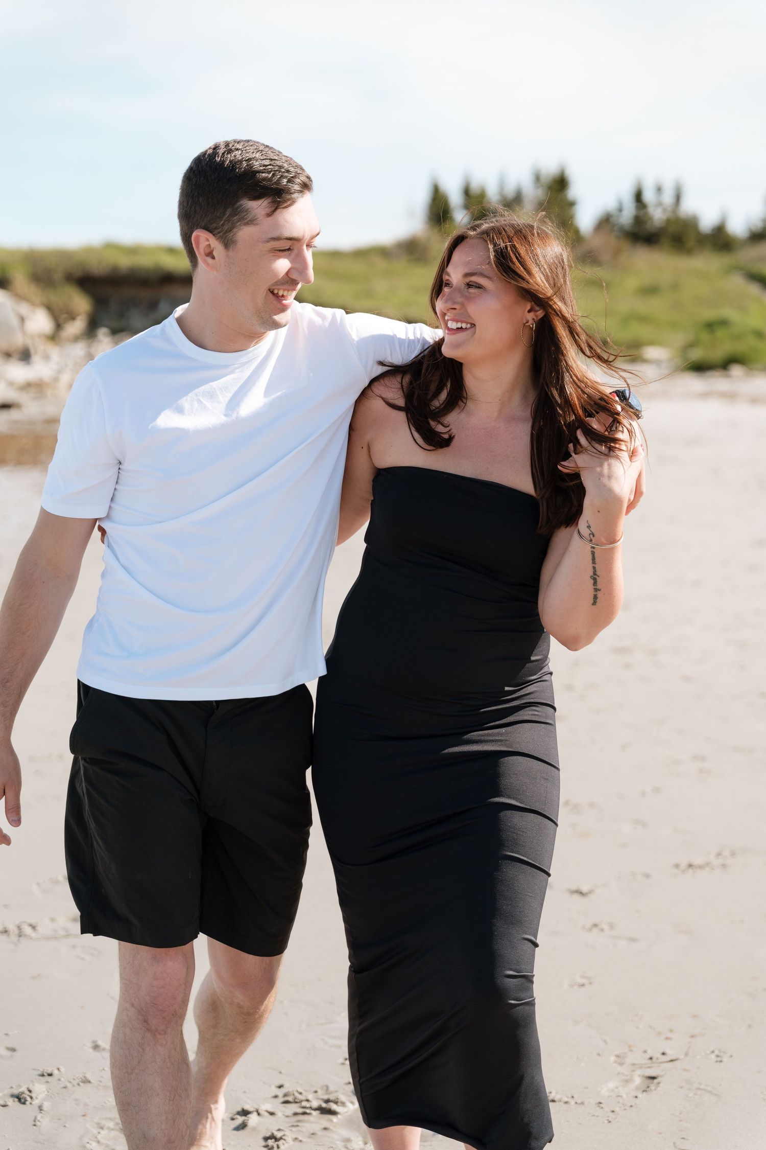 A happy couple walks on a sandy beach, man in white tee and black shorts, woman in black strapless dress.