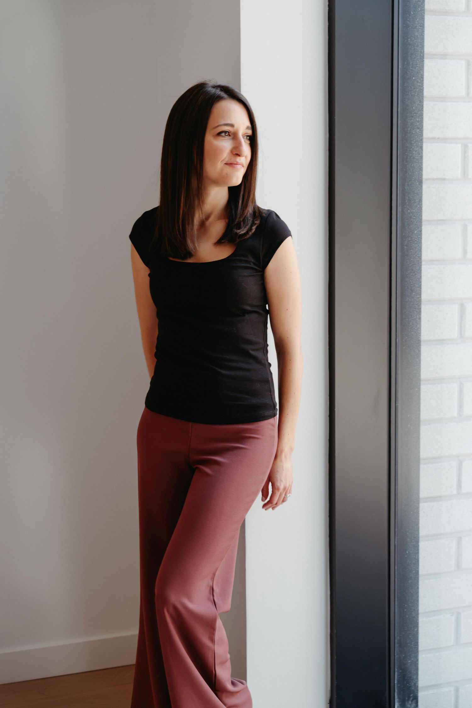 Woman in black top and mauve wide-leg pants standing by a window in a modern interior.