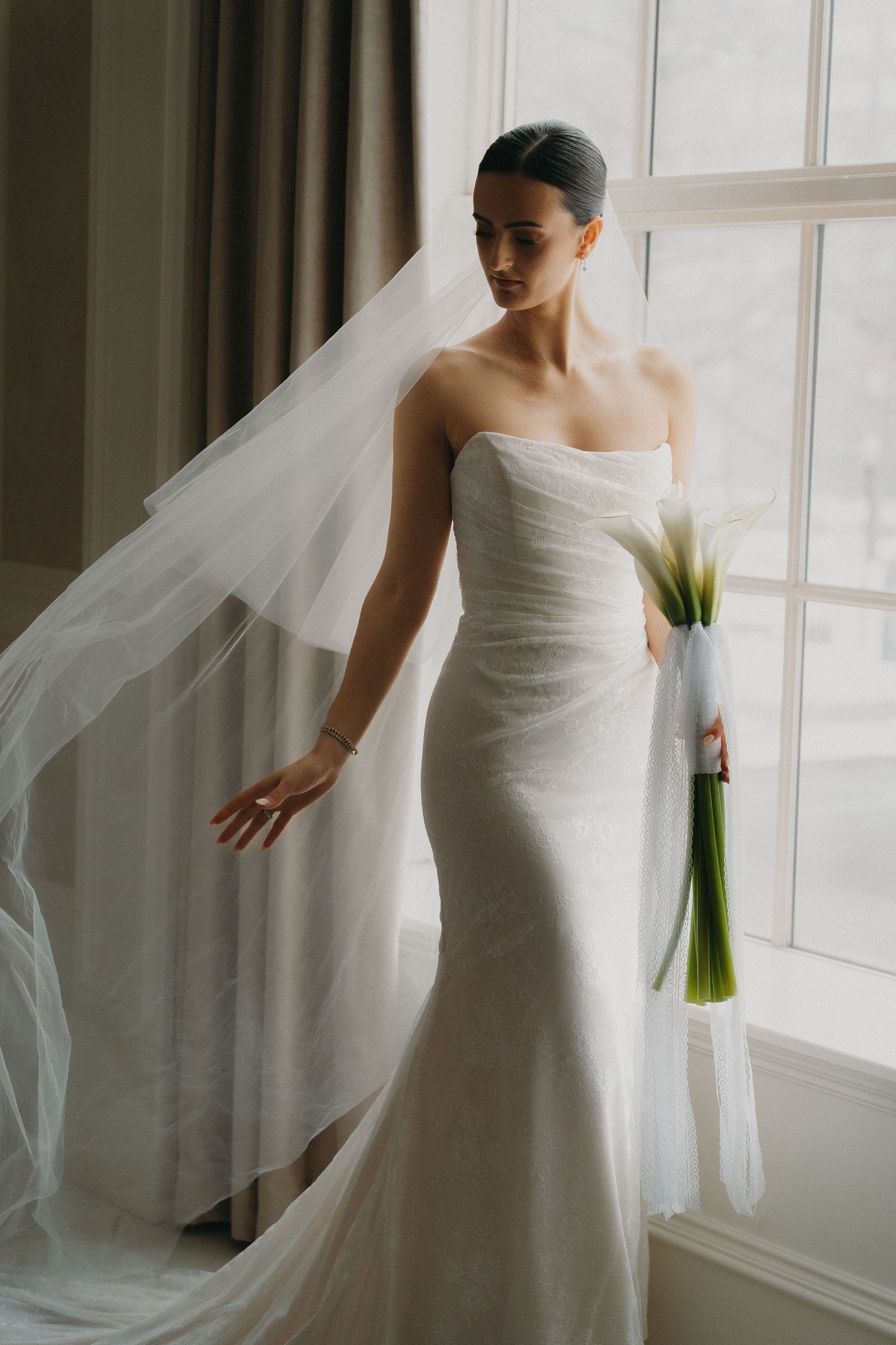 Elegant bride in strapless white mermaid wedding gown with flowing veil holds calla lily bouquet by window.