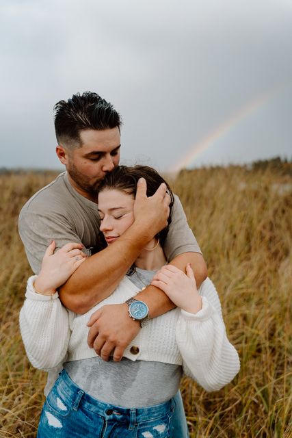 Couple embracing in golden field with rainbow visible in background.