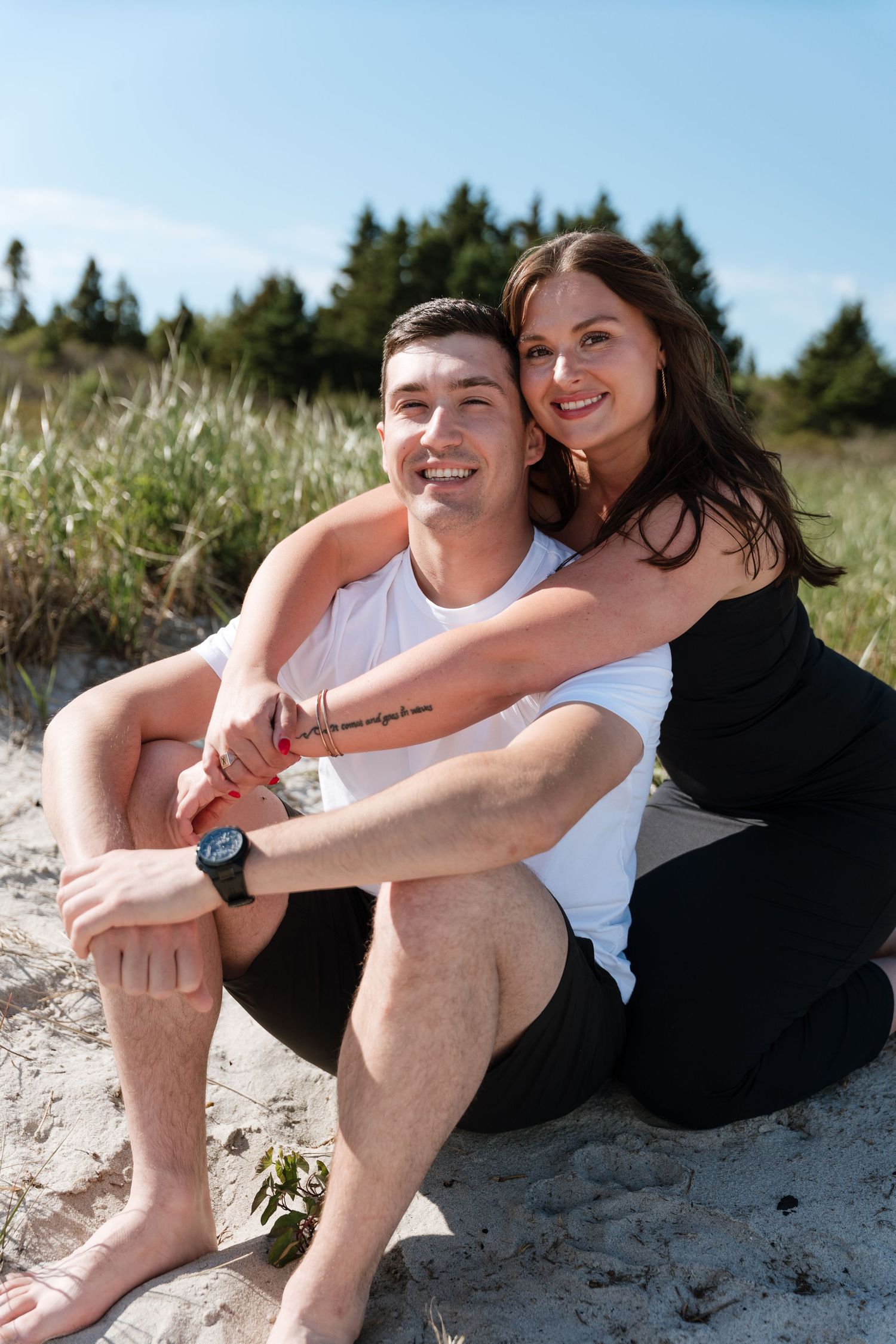 A happy couple sits together on a sandy beach, smiling, with trees and blue sky in the background.