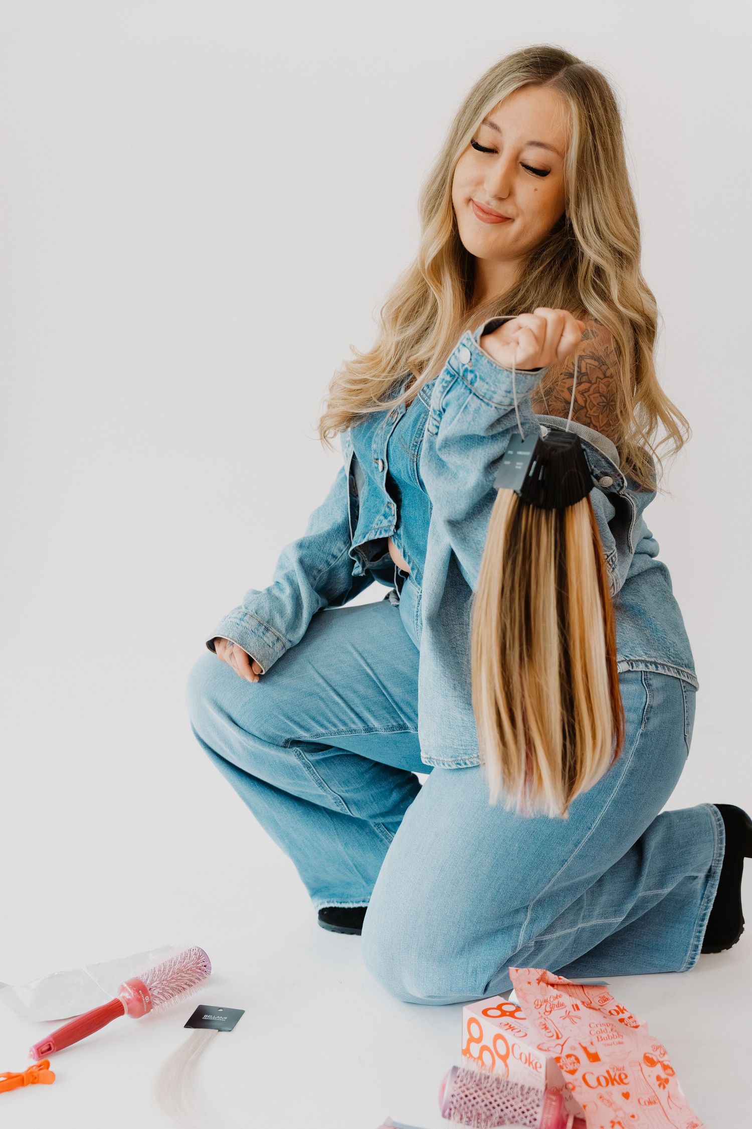 Blonde woman in denim outfit sitting on floor holding hair extensions with styling tools nearby.