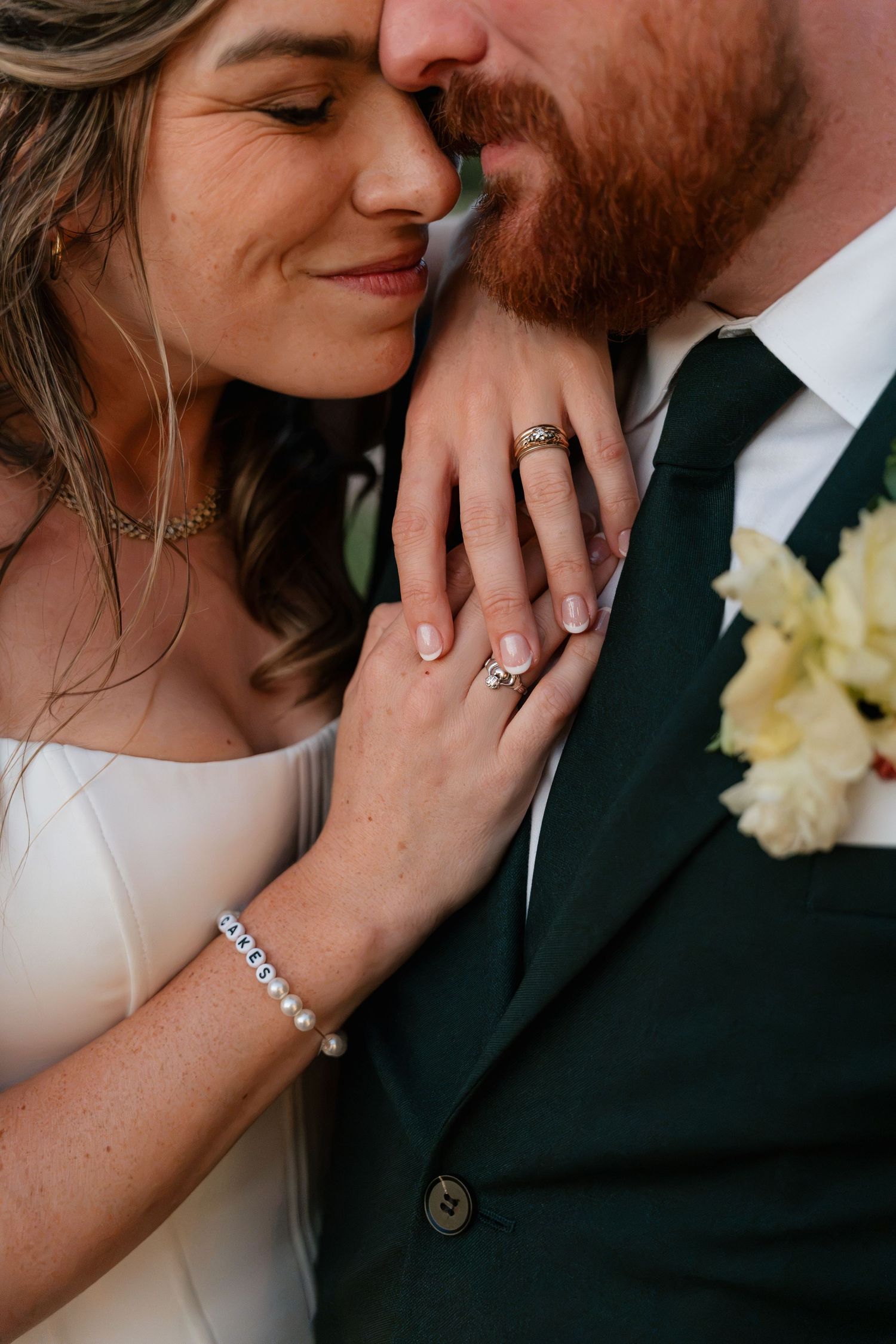Bride and groom closeup showing wedding rings, pearl bracelet, green suit with boutonniere, and tender romantic pose.